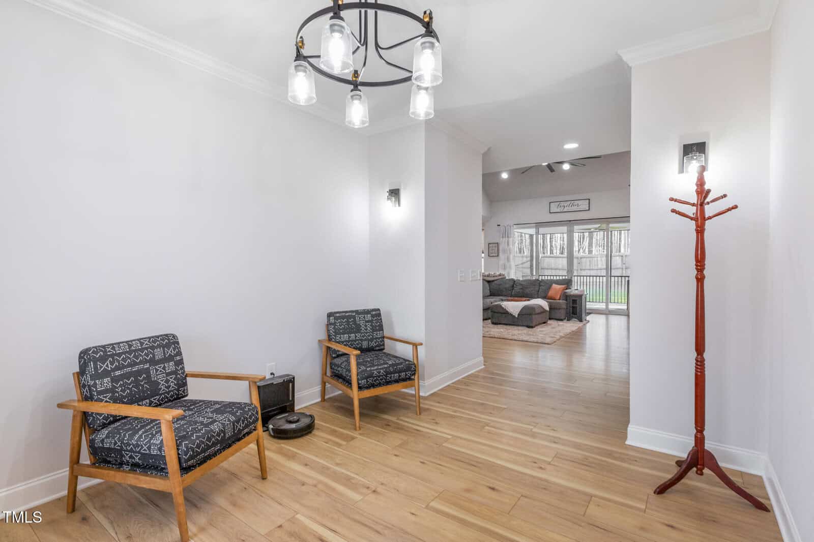 A bright hallway with light wood flooring, two patterned armchairs, a modern ceiling light, wall sconces, a red coat rack, and a view into a cozy living room with a sectional sofa and large windows.