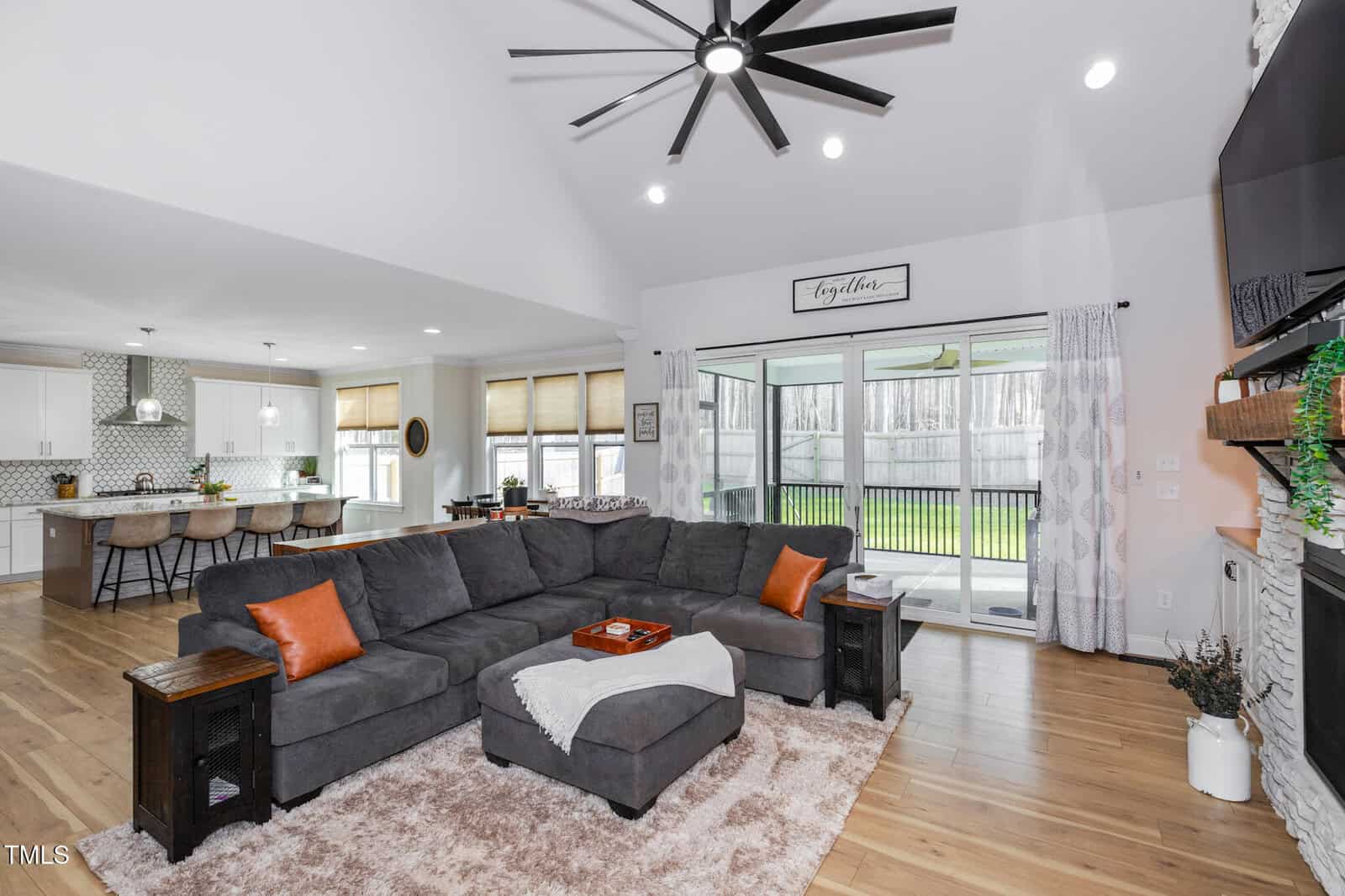 A spacious open-concept living room and kitchen featuring a large gray sectional sofa with orange pillows, a soft rug, a wall-mounted TV, a fireplace, and sliding glass doors opening to a fenced backyard.