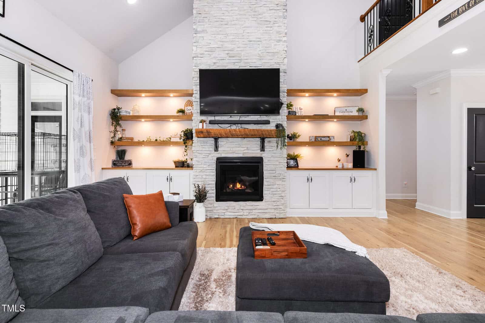 A modern living room with a gray sectional sofa, a stone fireplace with a mounted TV, built-in shelves with decorative items, and light wood flooring. A soft rug and ottoman with a tray are in front of the couch.