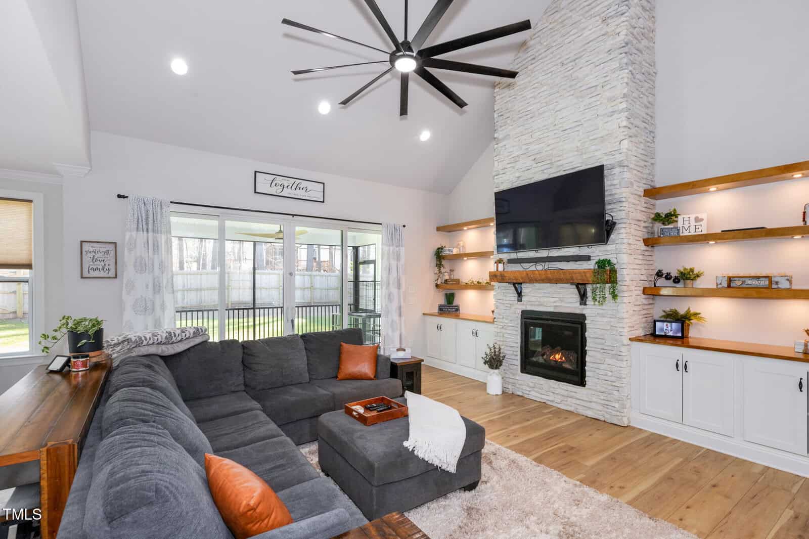 A modern living room with a tall stone fireplace, wall-mounted TV, built-in shelves with decor, large gray sectional sofa, wood flooring, and floor-to-ceiling windows showing a fenced backyard.