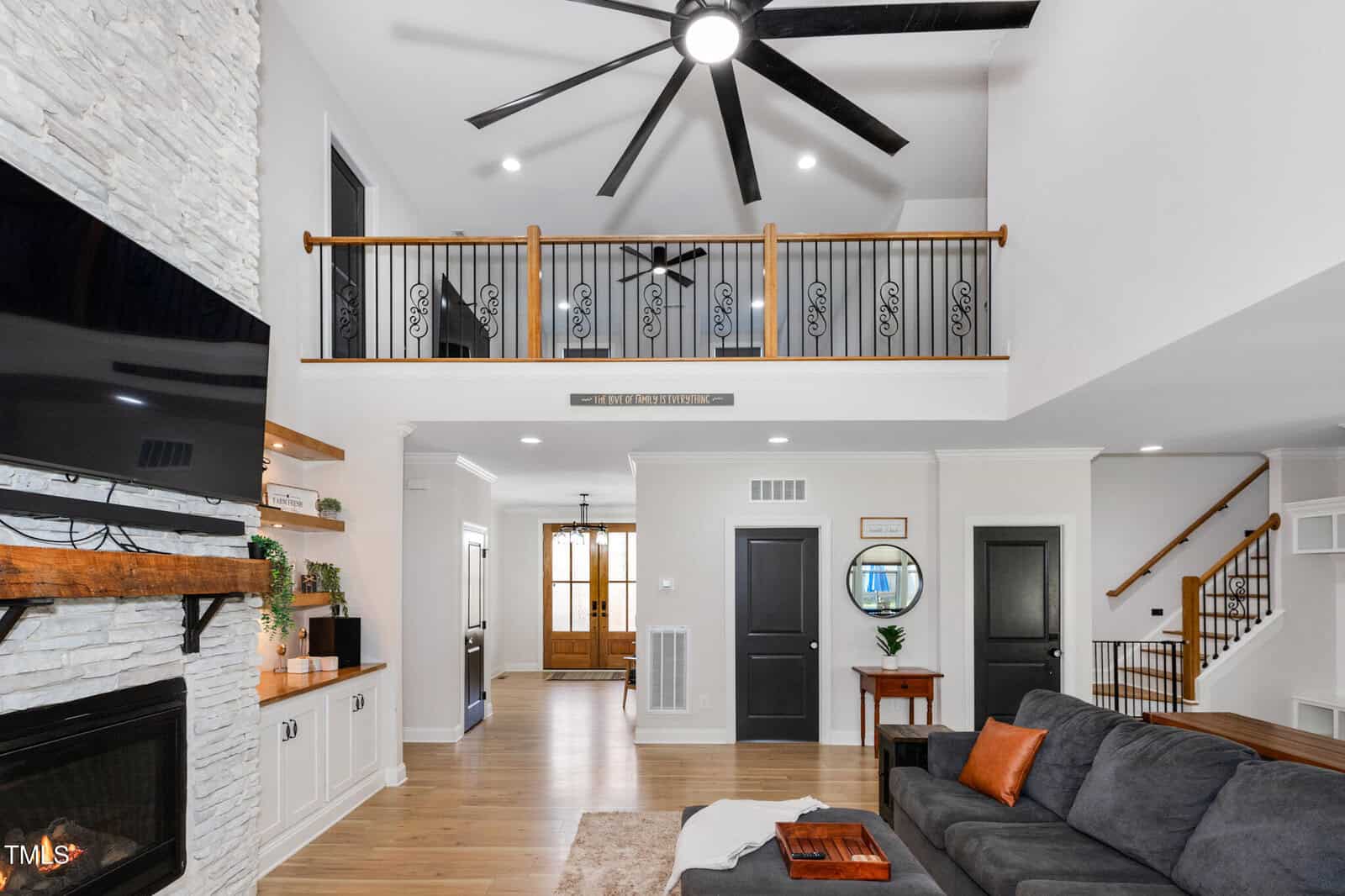 Modern living room with a gray sectional sofa, stone fireplace, wall-mounted TV, wood accents, and a loft with black railings. High ceiling features a large black fan; natural light fills the space.