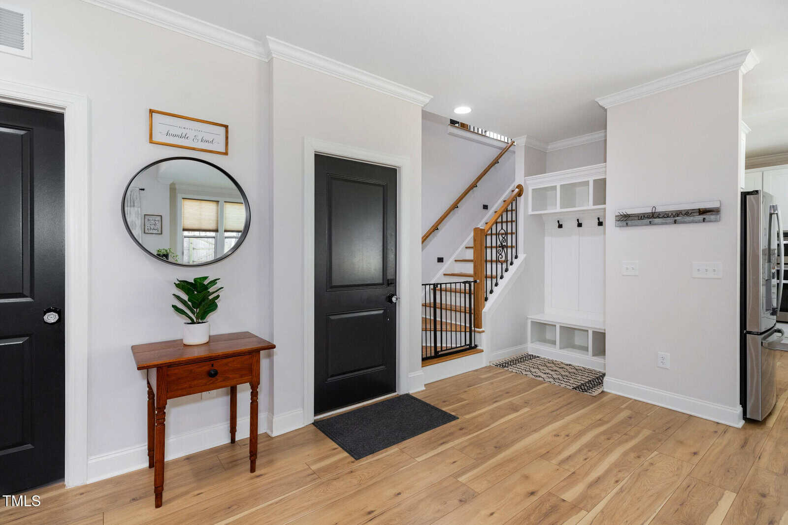 A bright entryway with wood floors, a small table with a plant, a round mirror, a black door, white walls, a staircase with a wooden handrail, and a built-in bench with hooks and cubbies.