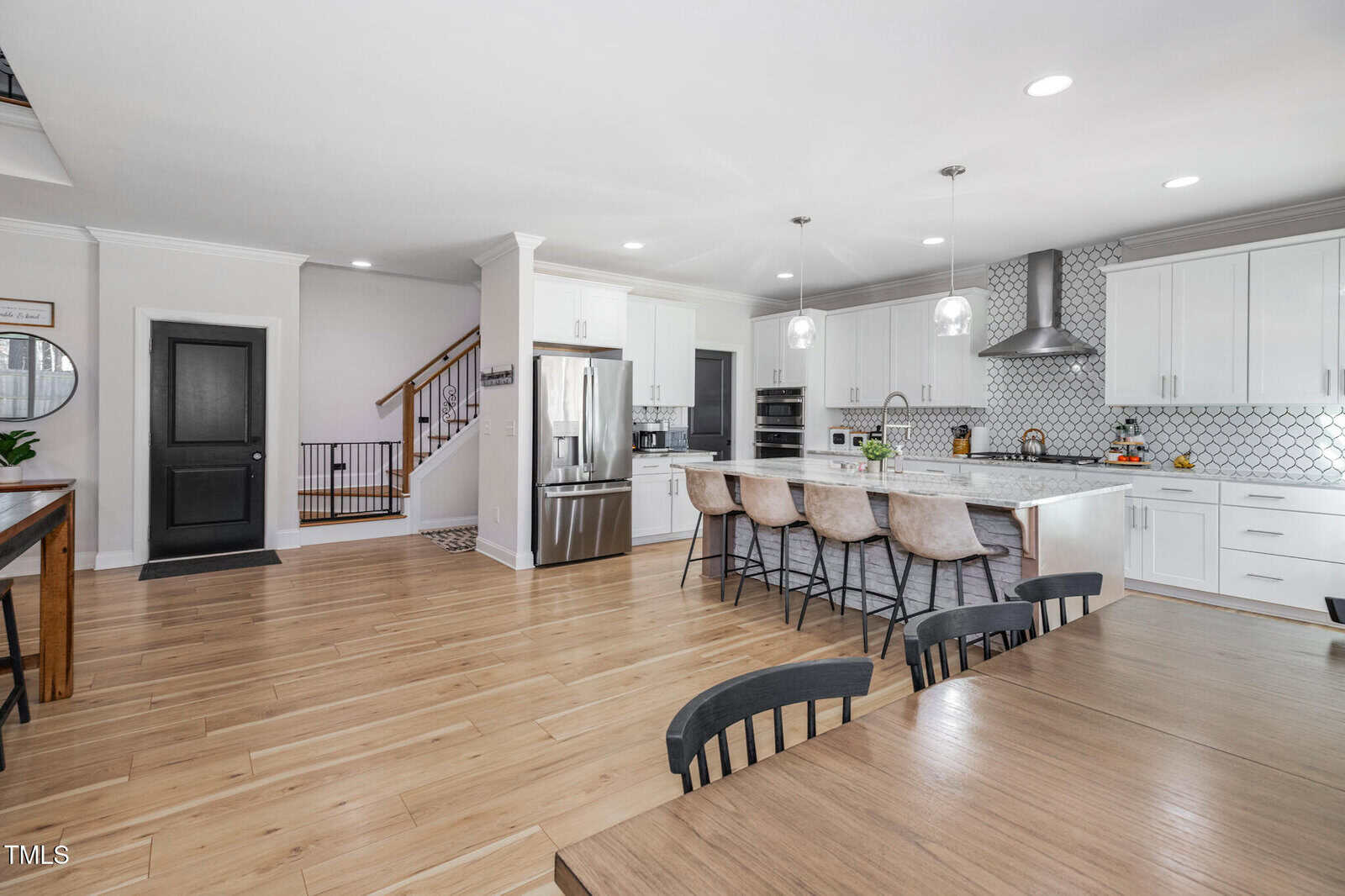 Spacious modern kitchen with white cabinets, a large island with barstools, stainless steel appliances, wood flooring, and a dining table in the foreground. Stairs and a black door are visible in the background.