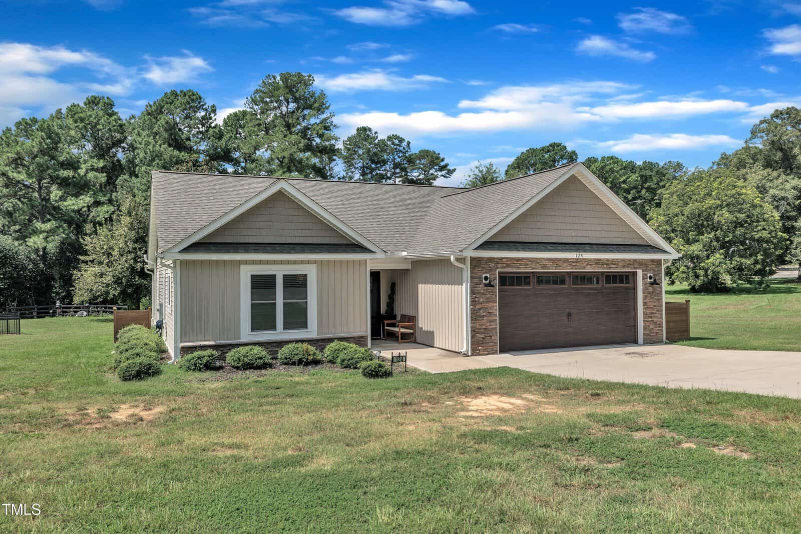 Single-story house with a front porch, stone and siding exterior, two-car garage, and well-kept lawn, surrounded by trees and under a blue sky with scattered clouds.