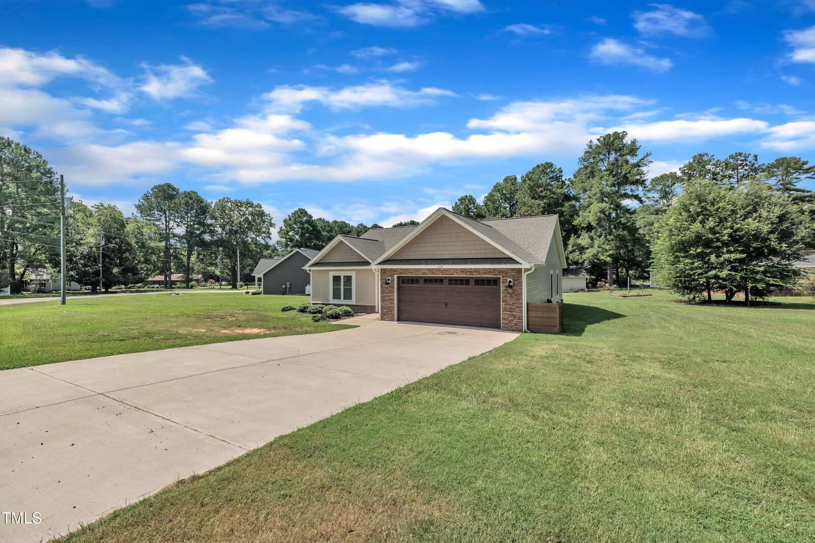 Single-story house with a brick and siding exterior, attached two-car garage, wide driveway, and a large grassy front yard, set against a backdrop of trees and a bright blue sky with scattered clouds.
