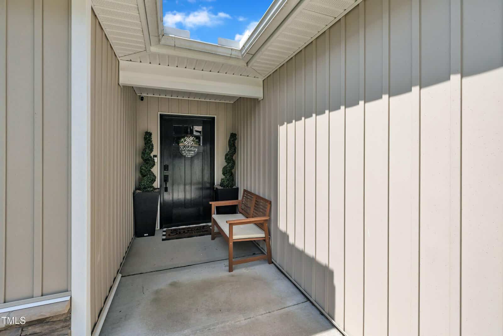 A covered entryway with beige vertical siding, a black front door with a welcome wreath, two potted topiary plants, and a wooden bench along the wall. The area is well-lit by a skylight above.