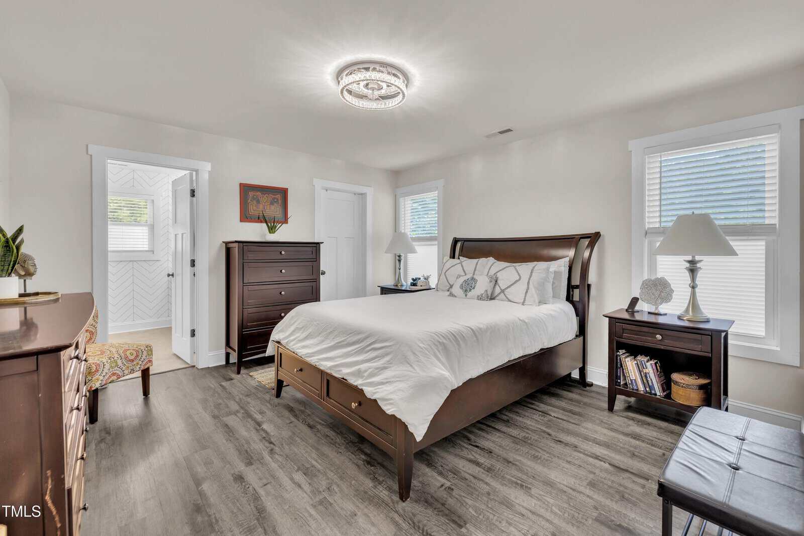 Bright bedroom with wood flooring, a dark wood bed frame, matching dresser and chest, white bedding, two windows with blinds, a chandelier ceiling light, and a small reading nook with a patterned chair and side table.