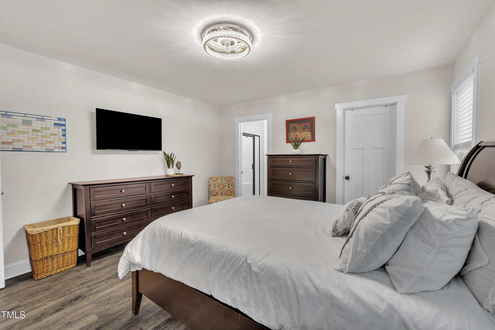 A bright bedroom with a bed, white bedding, dark wood furniture, a wall-mounted TV, wicker laundry basket, calendar, and modern light fixture. Decorative pillows and framed art add touches of color.
