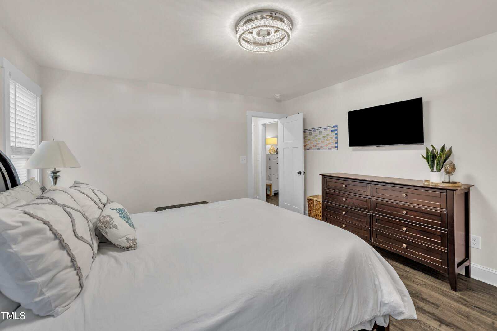 A bedroom with a neatly made bed, white walls, a ceiling light, a flat-screen TV on the wall, a dark wooden dresser with decor, and an open door leading to another room.