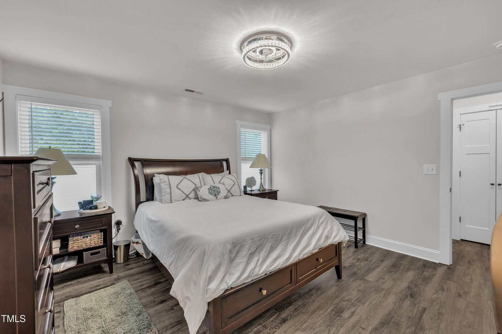 A bedroom with a wooden bed frame, white bedding, two matching bedside tables with lamps, a dresser, a small bench at the foot of the bed, wood flooring, and a modern ceiling light fixture.
