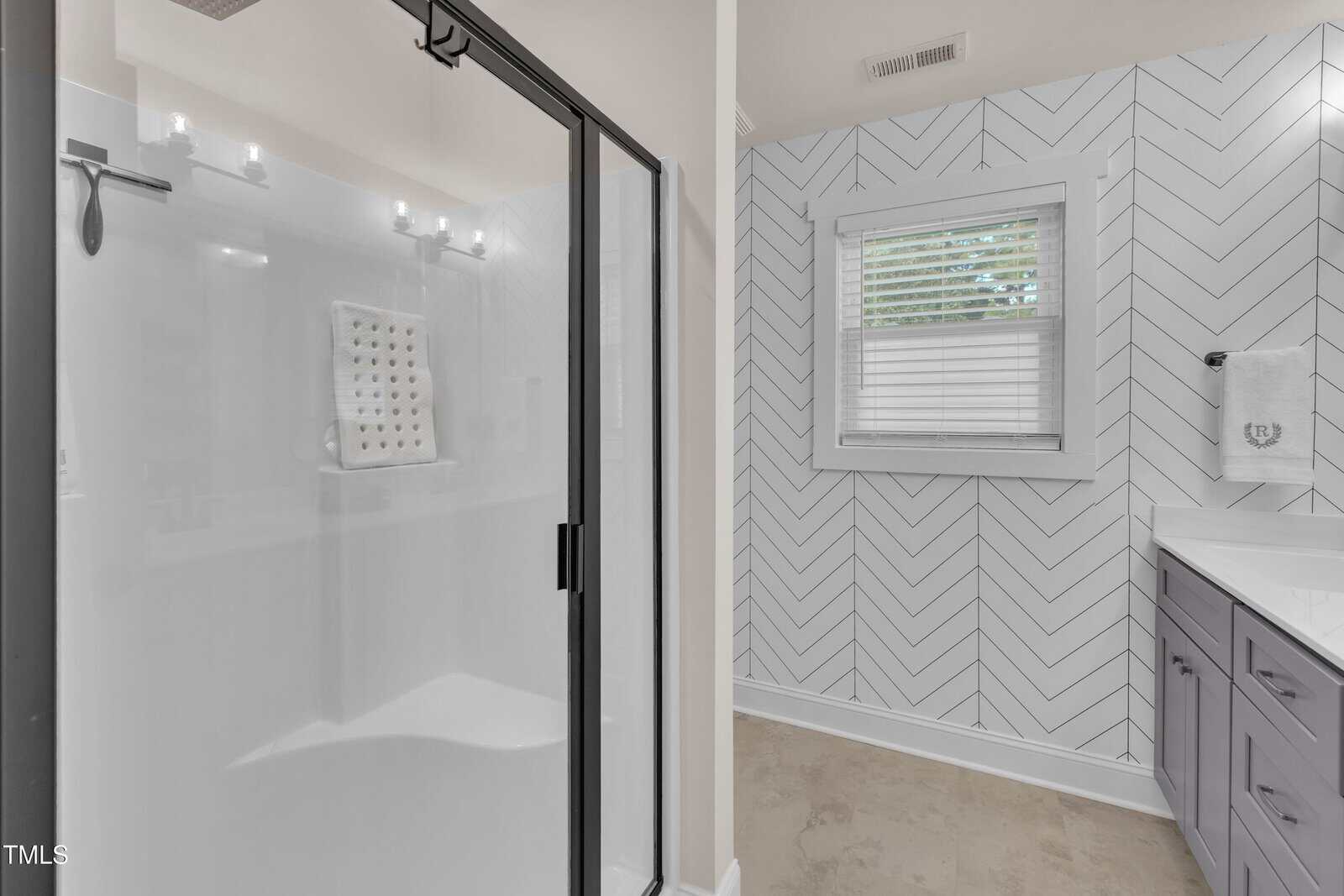 Modern bathroom with a glass-enclosed shower, a vanity with drawers, herringbone-patterned white tile walls, a window with white blinds, and a towel hanging beside the sink.