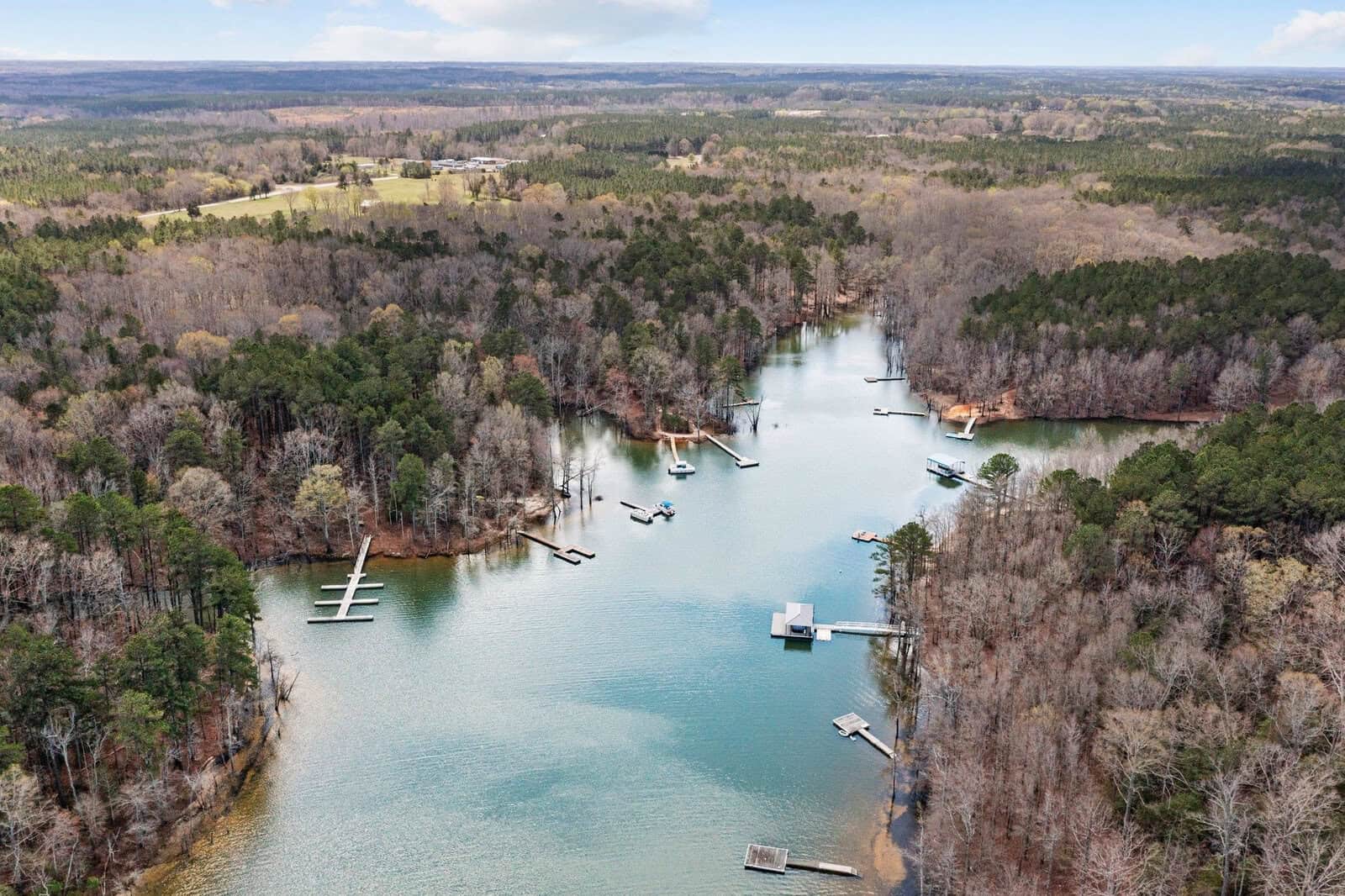 Aerial view of a wooded lakeshore with several docks extending into calm water, surrounded by dense trees and forest. A few boats are moored at the docks. The sky is partly cloudy.