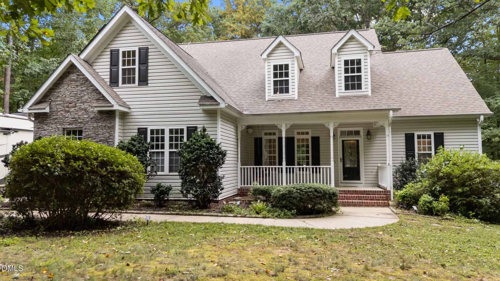A light gray house with dormer windows, a stone accent wall, white trim, and a covered front porch with columns sits gracefully at 395 Timberlands Drive in Louisburg. There are bushes and a lawn in front, with trees in the background.