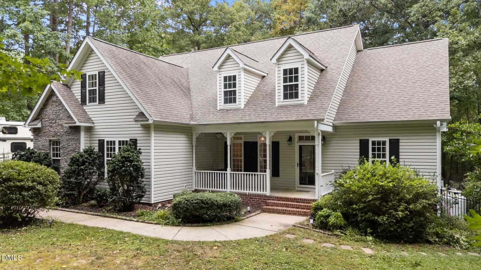 A two-story house with beige siding, multiple peaked roofs, black shutters, and a covered front porch. There are bushes and trees surrounding the house and a curved concrete walkway leading to the front steps.