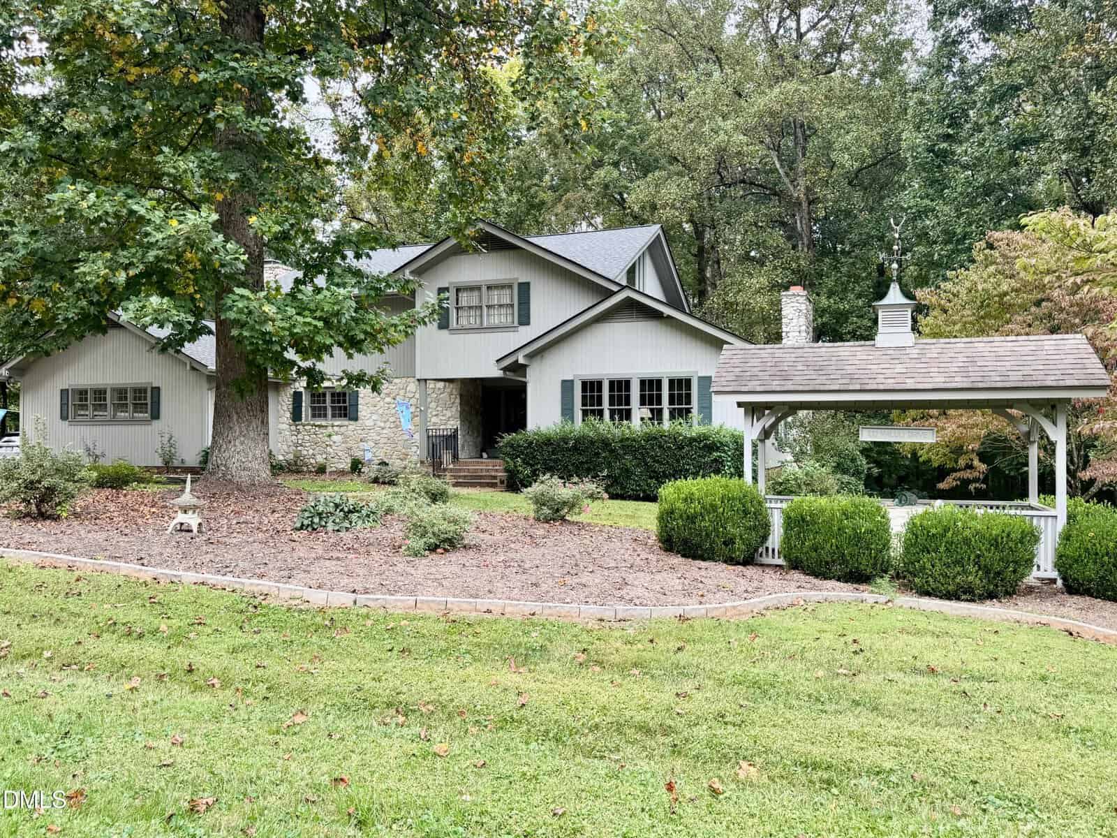 A gray two-story house with stone accents sits on a sloped lawn at 100 Maluli Drive, Oxford, surrounded by trees and bushes. A small white gazebo-style structure with a weathervane is on the right side of the property.