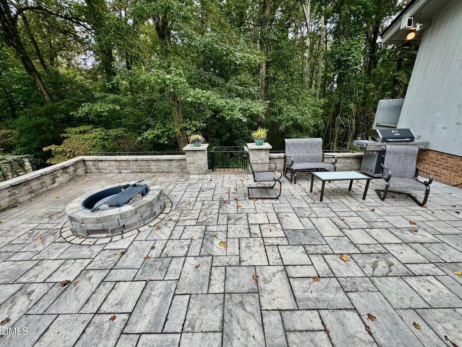 Stone patio with a built-in fire pit, outdoor furniture including chairs and a grill, bordered by a low stone wall and surrounded by dense trees in the background.