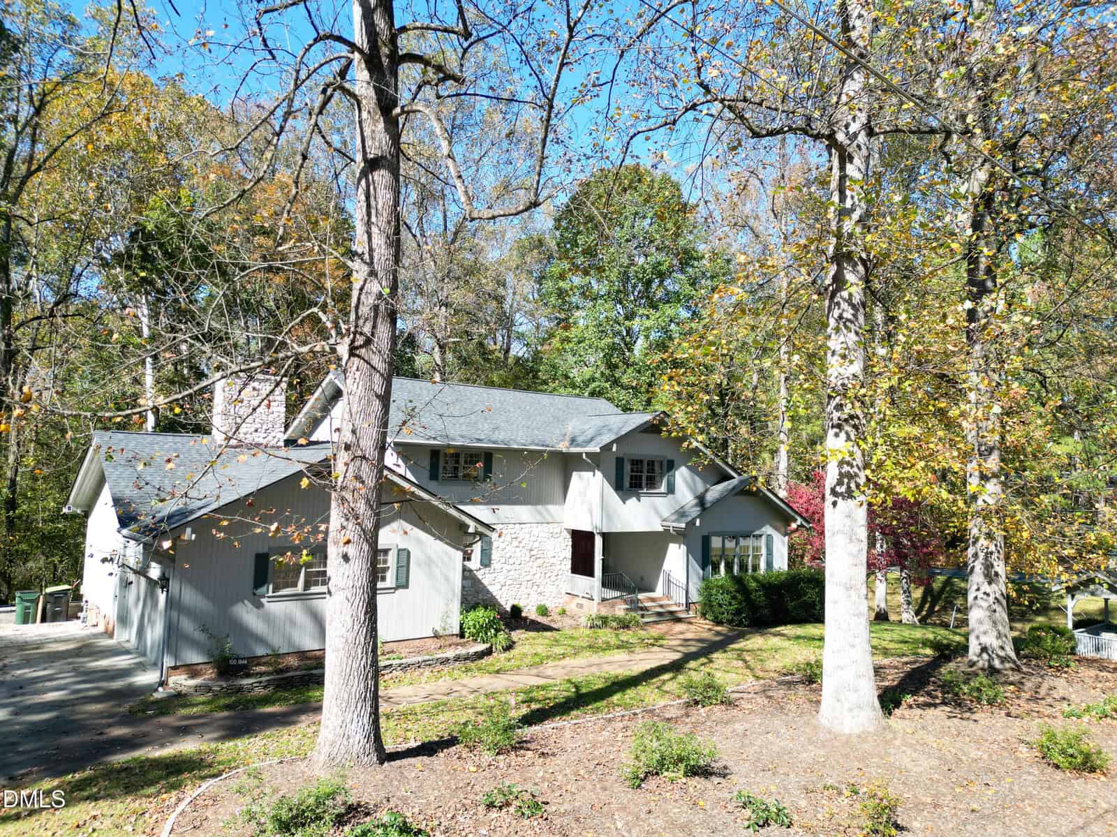Two-story house with gray siding and stone accents, set among tall trees with autumn leaves. A paved driveway curves to the left, and the house sits in a wooded area with sunlight filtering through the branches.