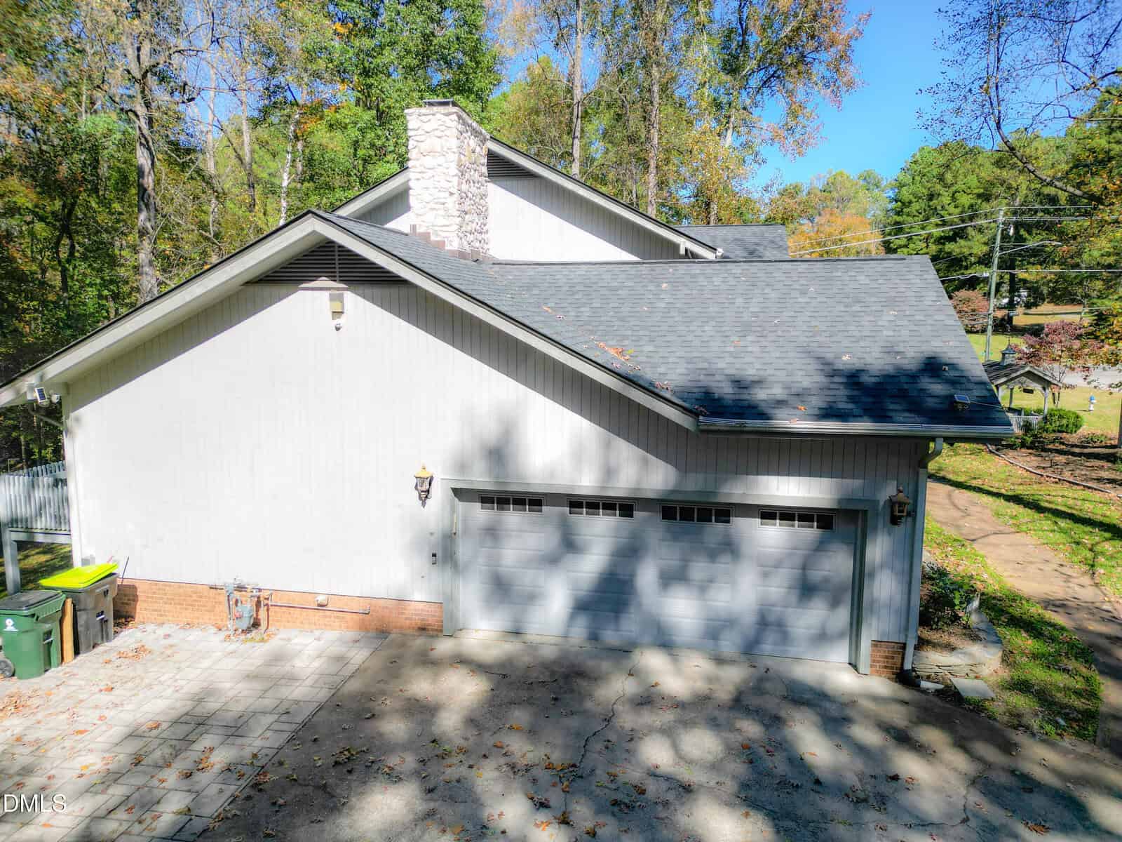 A two-car garage with white doors attached to a light gray house with a brick foundation, stone chimney, and sloped roof; surrounded by trees and a paved driveway.