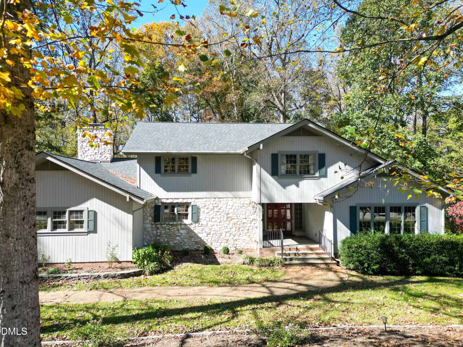 A two-story house with light gray siding, stone accents, and a dark gray roof, surrounded by trees and a front yard with a walkway and shrubs on a sunny day.