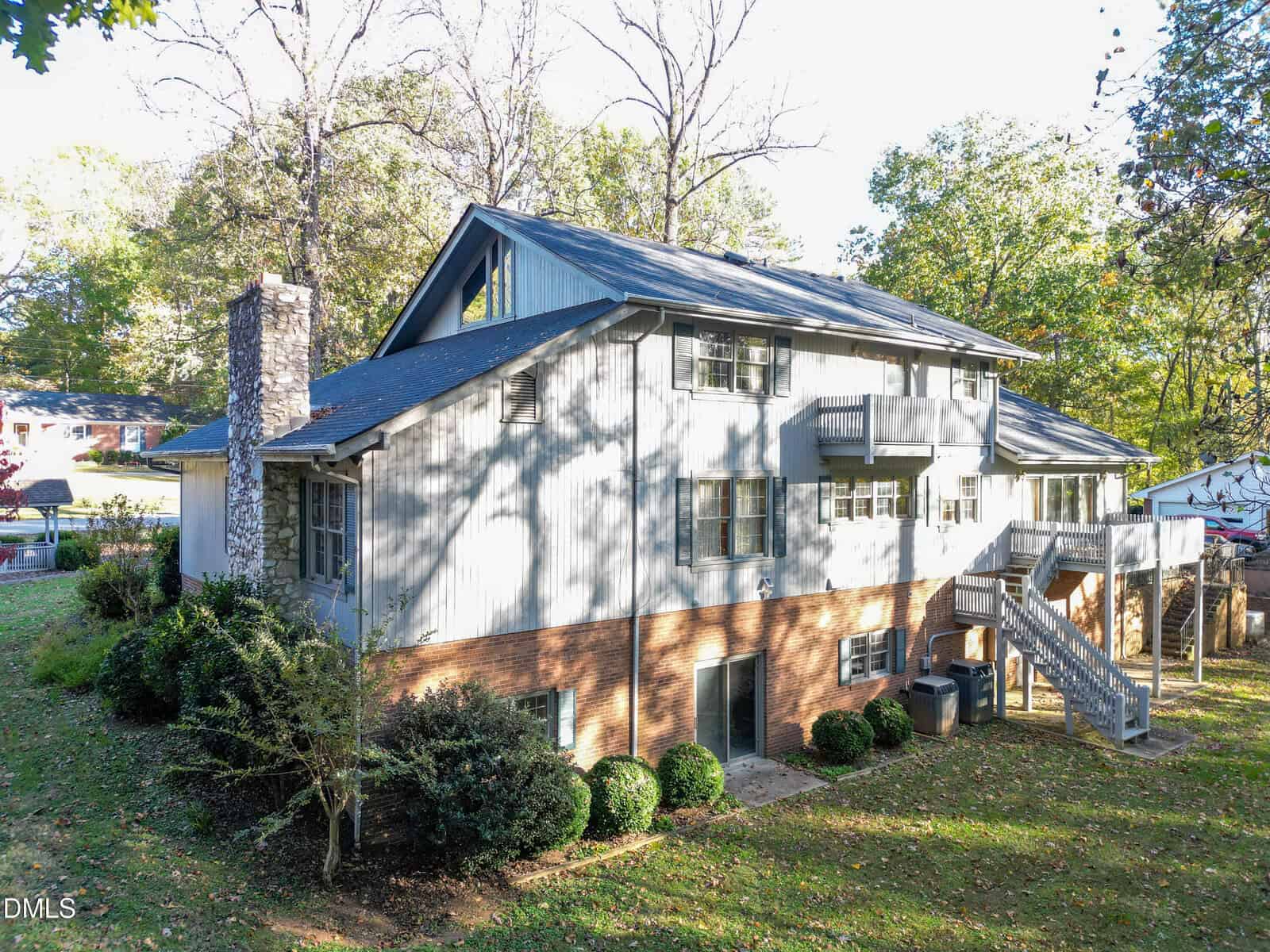 Two-story house with light gray siding, brick foundation, and a stone chimney. The home features multiple windows, a small balcony, an exterior staircase, and is surrounded by trees and lawn.