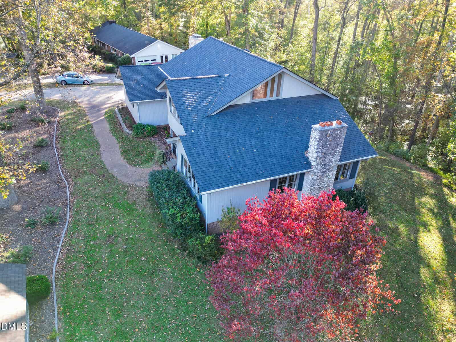 Aerial view of a two-story house with a dark blue roof, prominent stone chimney, and light-colored siding, surrounded by trees and a curved driveway, with a detached garage and car visible in the background.