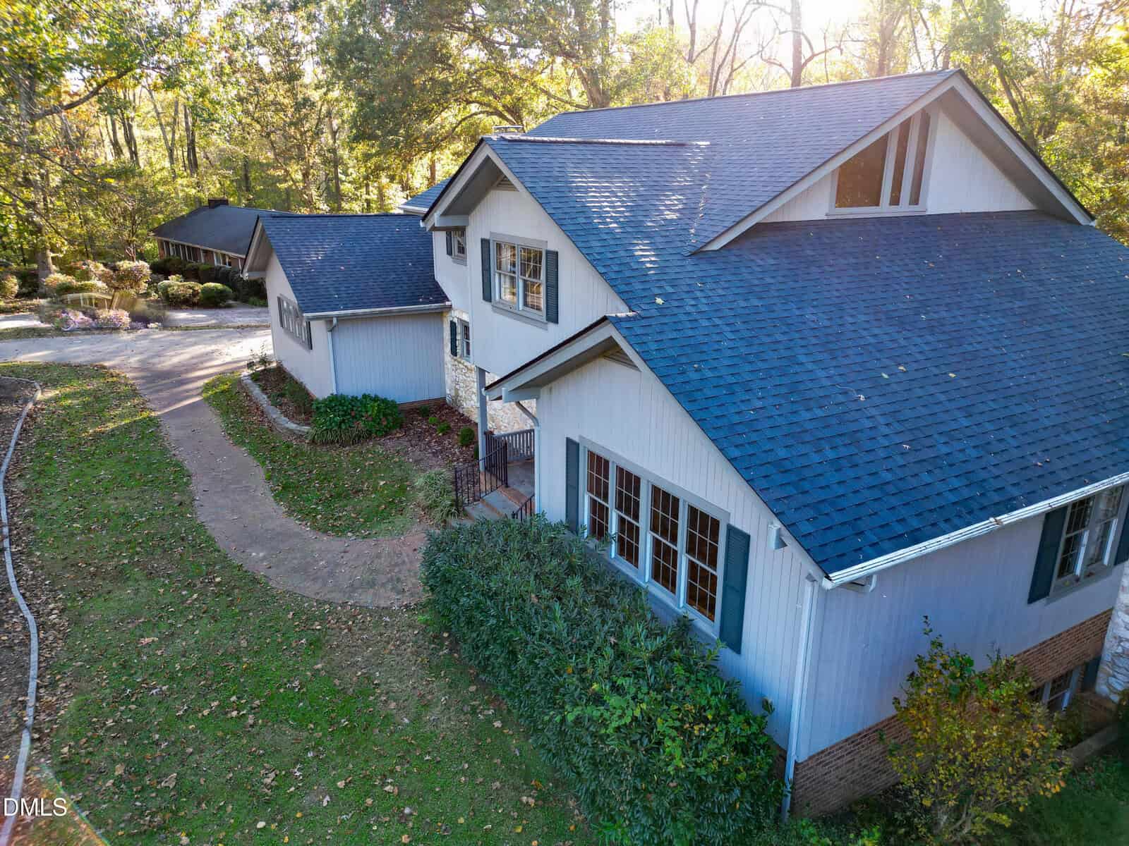 A two-story light blue house with dark roof shingles, white trim, and black shutters, surrounded by trees and landscaping with a curved walkway leading to the entrance. Sunlight filters through the trees in the background.