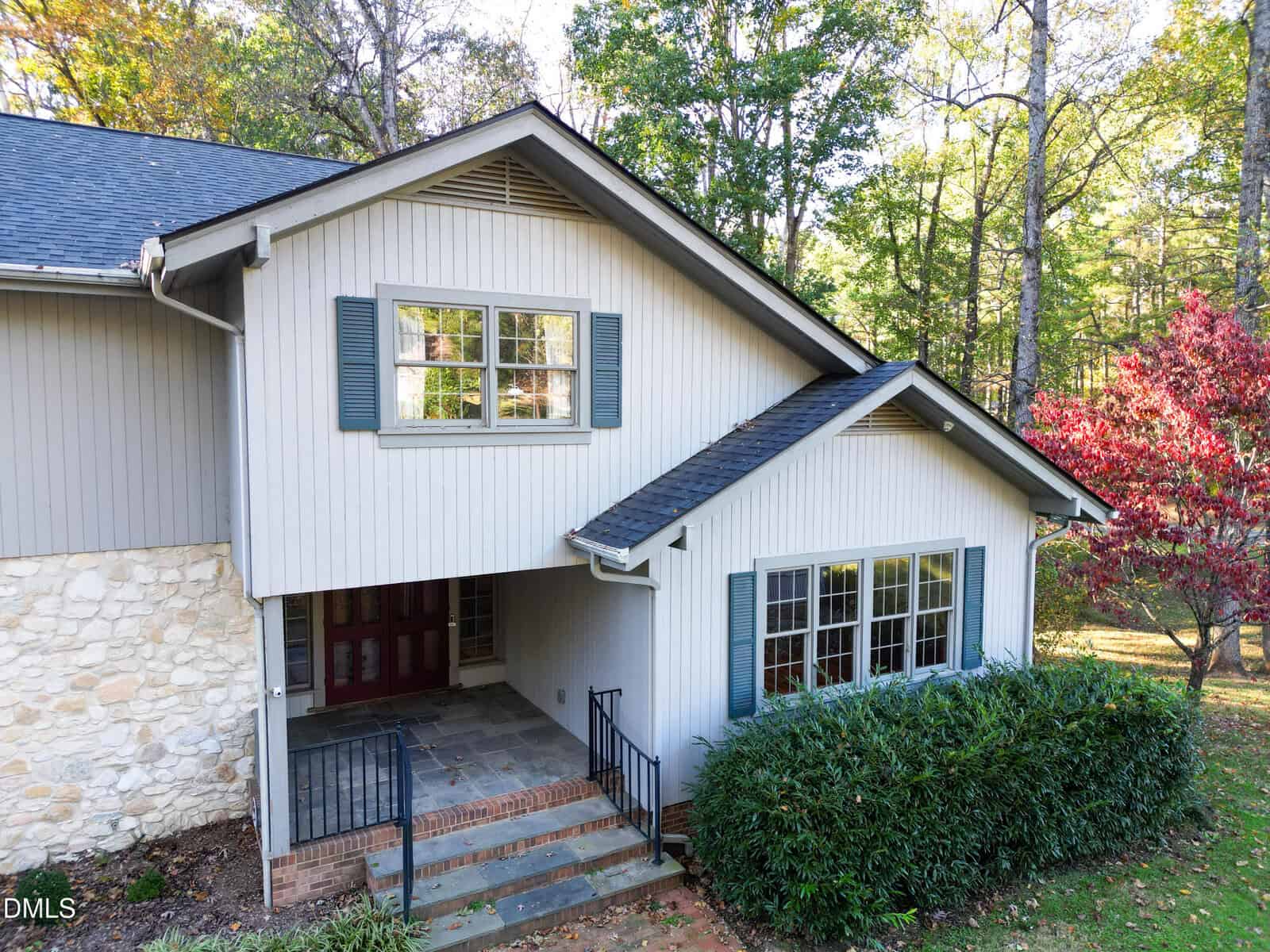 A two-story house with light gray siding, blue shutters, and a partial stone facade. There is a covered entryway, a brick step, metal railings, large front windows, and shrubs in front. Trees with autumn leaves are in the background.