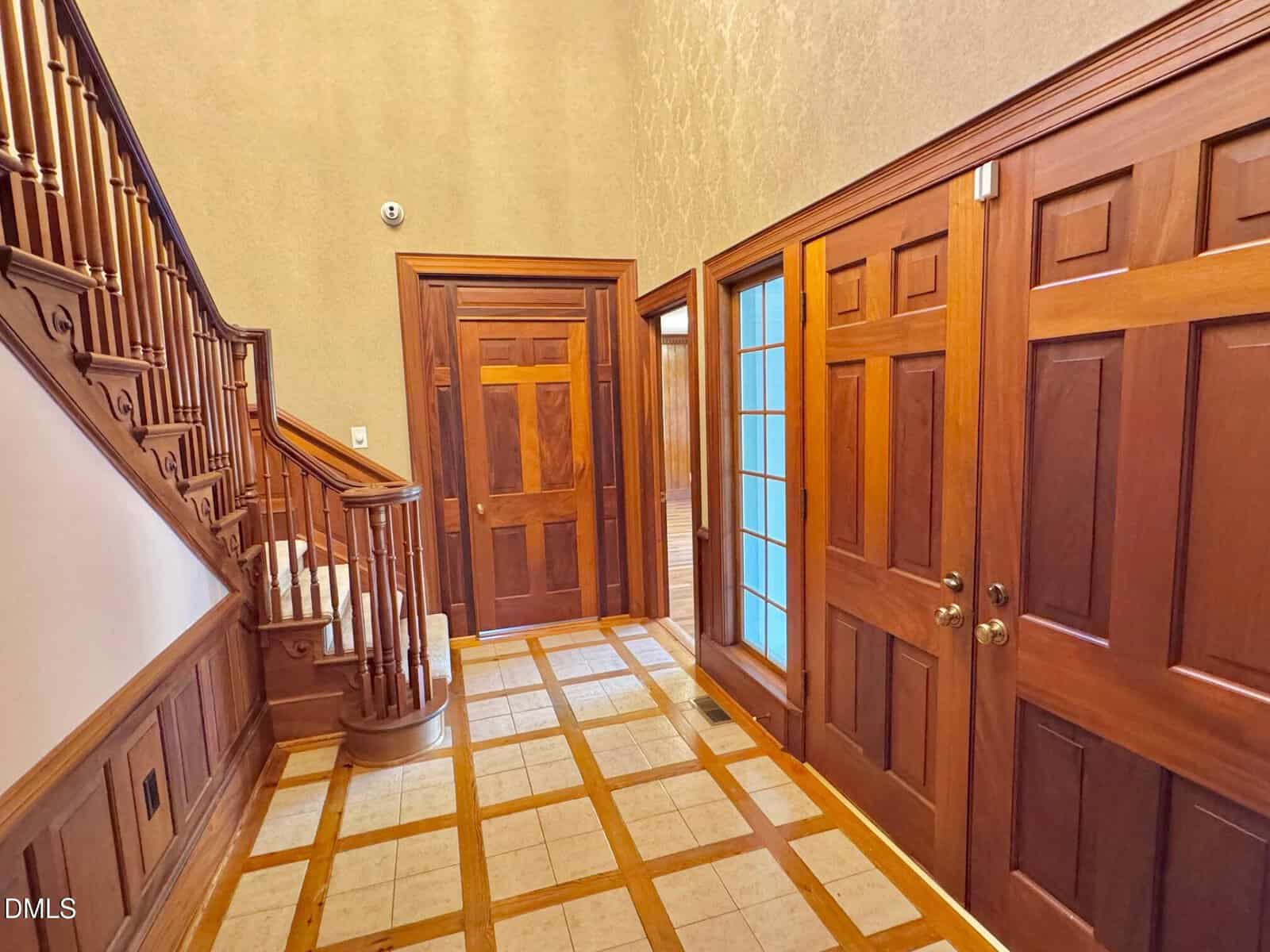 A foyer with wooden paneling, two closed wooden doors, a glass-paned door, and a staircase with wooden railings. The floor features a checkered tile pattern with wood trim. Walls are painted in a light neutral tone.