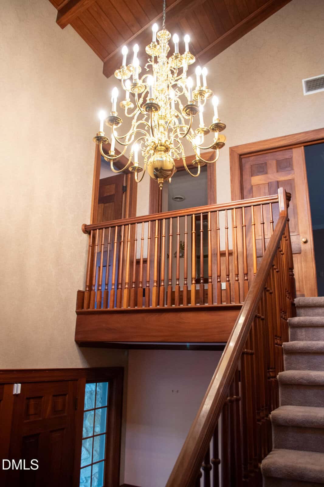 A gold chandelier with lit bulbs hangs above a wooden staircase and railing in a house with beige walls, wooden doors, and a carpeted stairway. Light from the chandelier brightens the entryway and upper hallway.