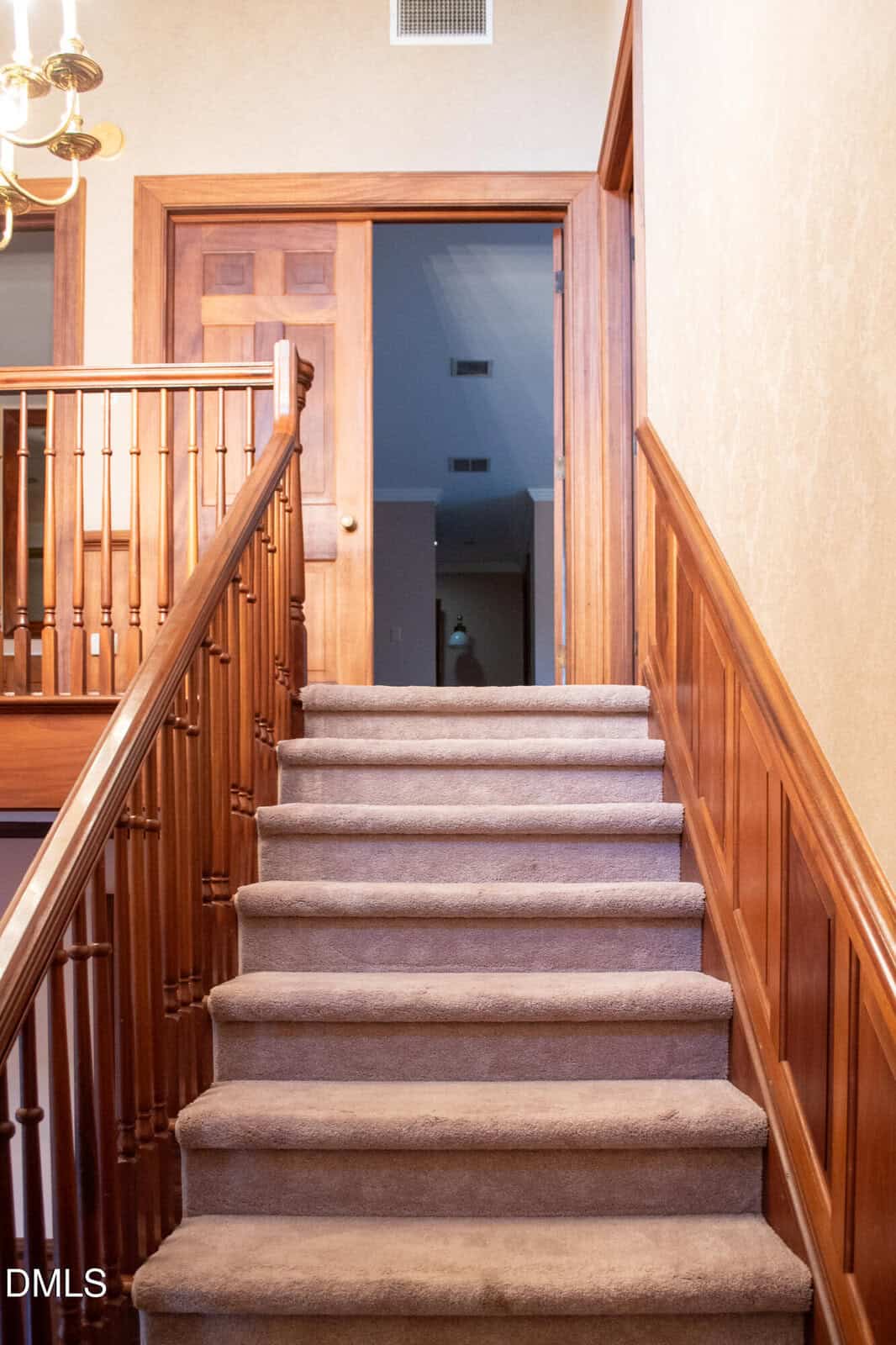 Carpeted staircase with wooden banister and wall paneling leads up to a landing with a partially open wooden door. Light fixture is visible at the top left. Warm, neutral tones throughout.