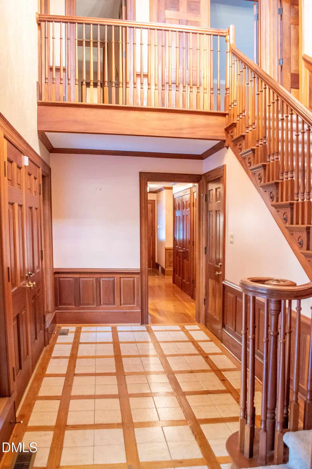 A foyer with tiled floors, wooden paneling, and a staircase leading to an upper level. There are multiple wooden doors and a hallway in the background. The space is well-lit with natural light.