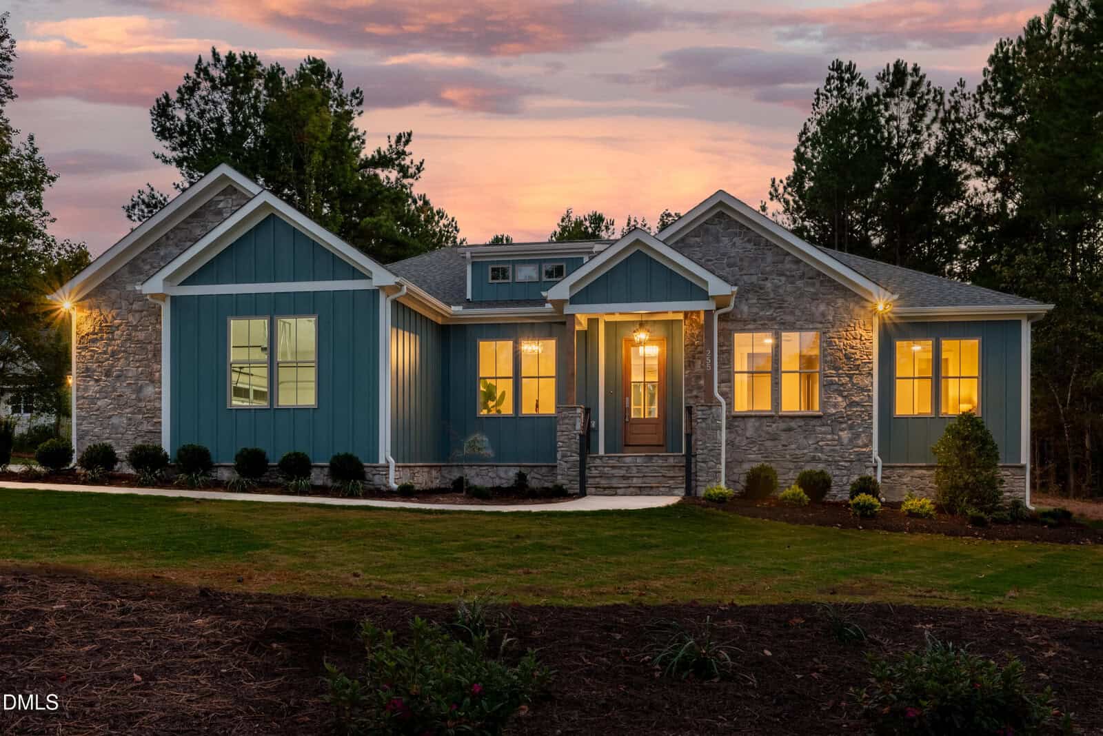 Single-story modern house with blue siding and stone exterior walls, large windows, a front porch with lights on, surrounded by trees and a landscaped yard at sunset, located at 255 Whistlers Cove in Louisburg.