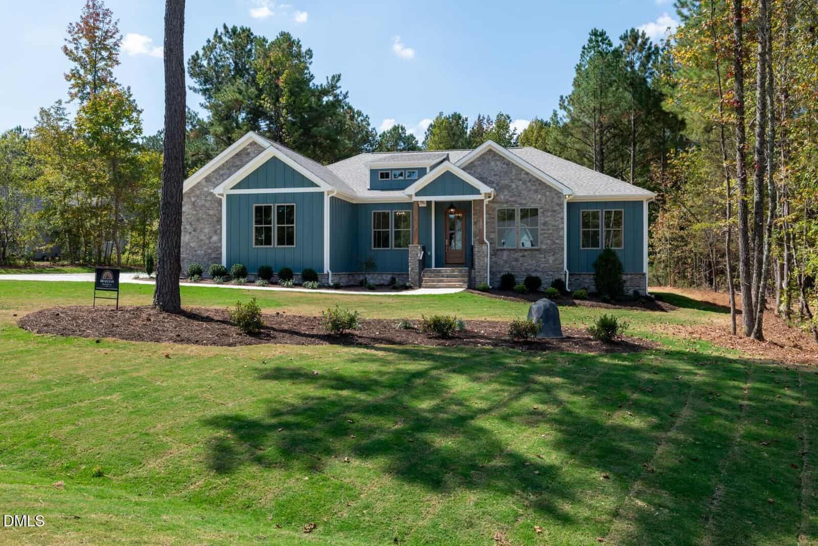 Single-story house with blue siding and stone accents, large windows, and a front porch. Surrounded by trees, the home sits on a well-maintained lawn with a curved driveway leading to the entrance.