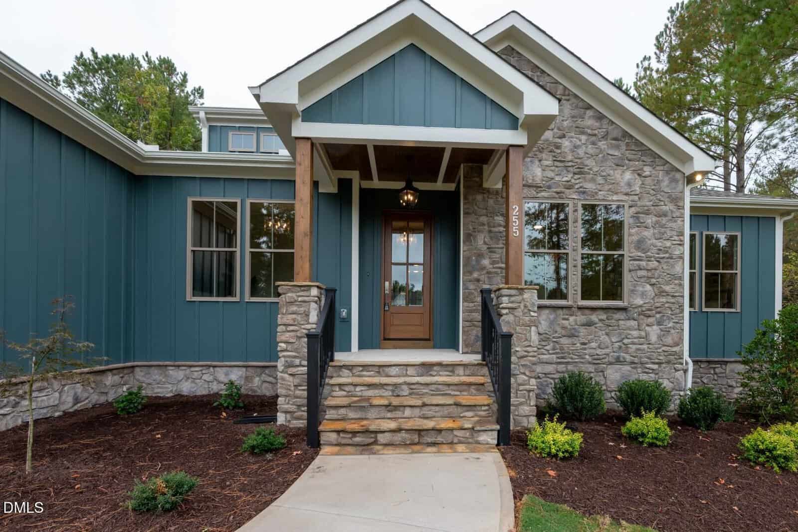 Front view of a modern house with a combination of stone and blue panel siding, a wooden front door with glass, a small covered porch with steps, and landscaped garden beds along the walkway.