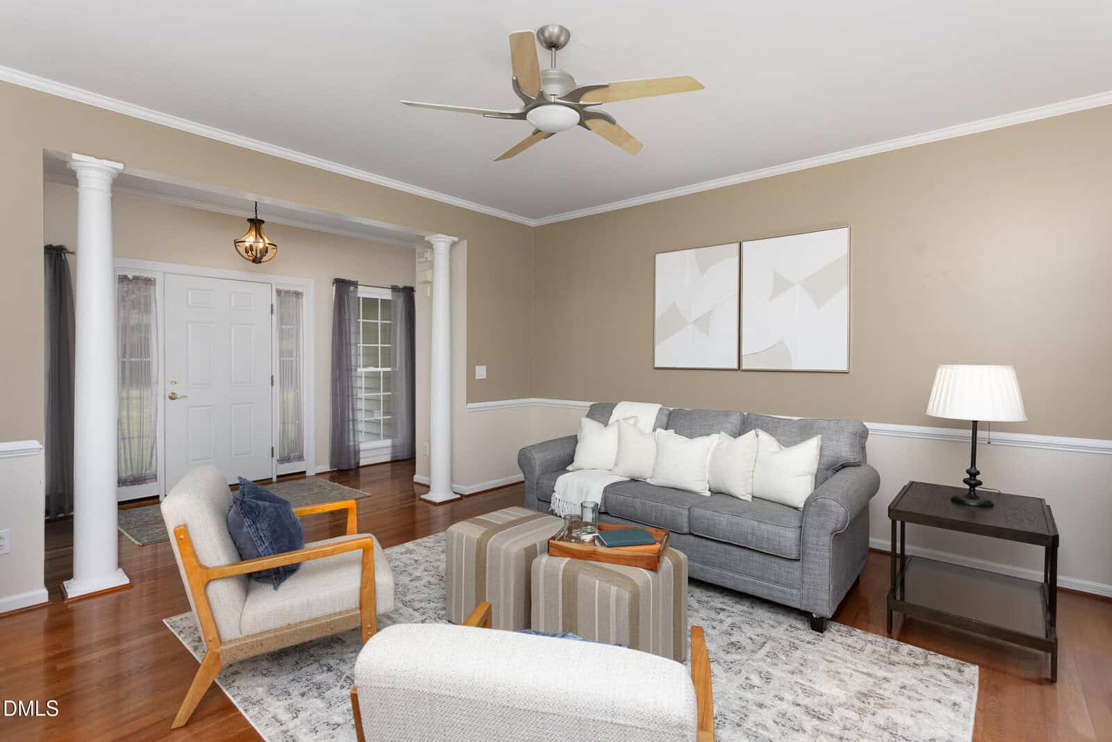 A living room with a gray sofa, two beige armchairs, a patterned ottoman, and a rug. A side table with a lamp is next to the sofa. The room has wood flooring, neutral walls, and white trim with columns.