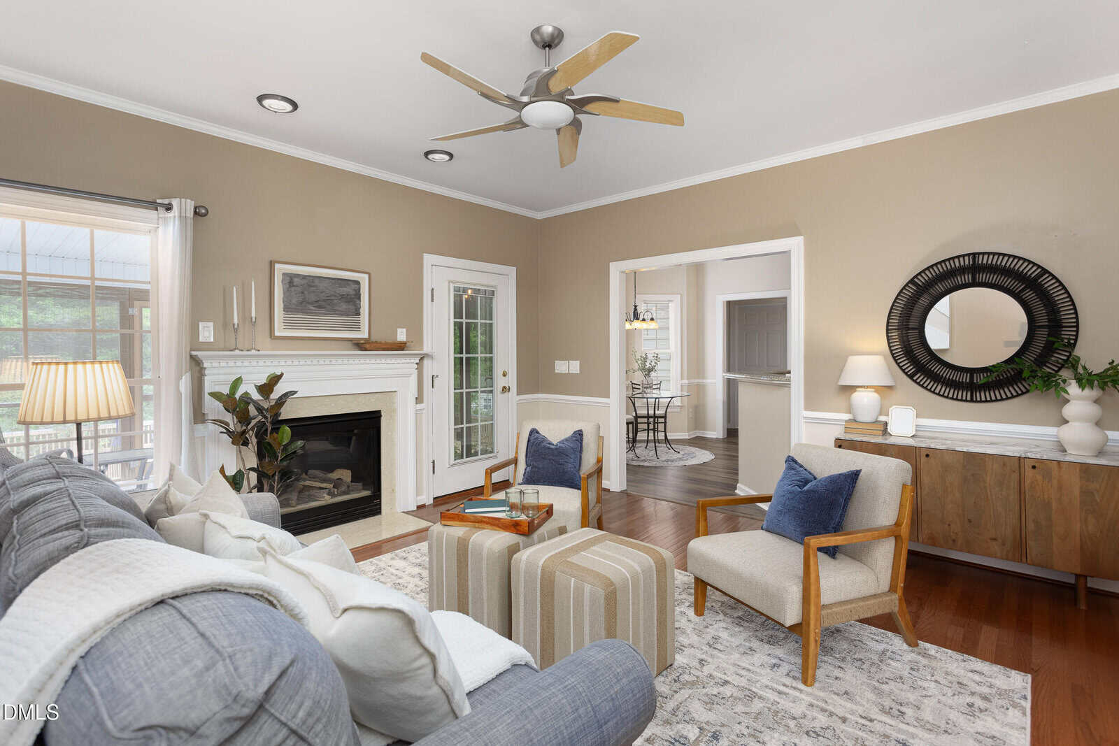 A living room with a sofa, two armchairs, a fireplace, a wooden ceiling fan, a sideboard with a mirror above it, and a door leading to another room. Neutral colors and natural light fill the space.