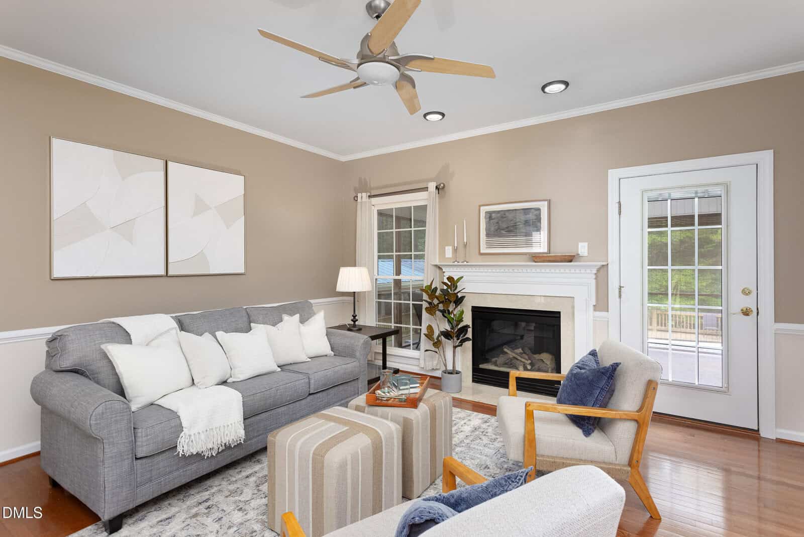 A living room with tan walls, a gray sofa, two armchairs, a white fireplace, modern abstract art, a ceiling fan, glass door, windows, wood floors, a rug, and decorative pillows and plants.