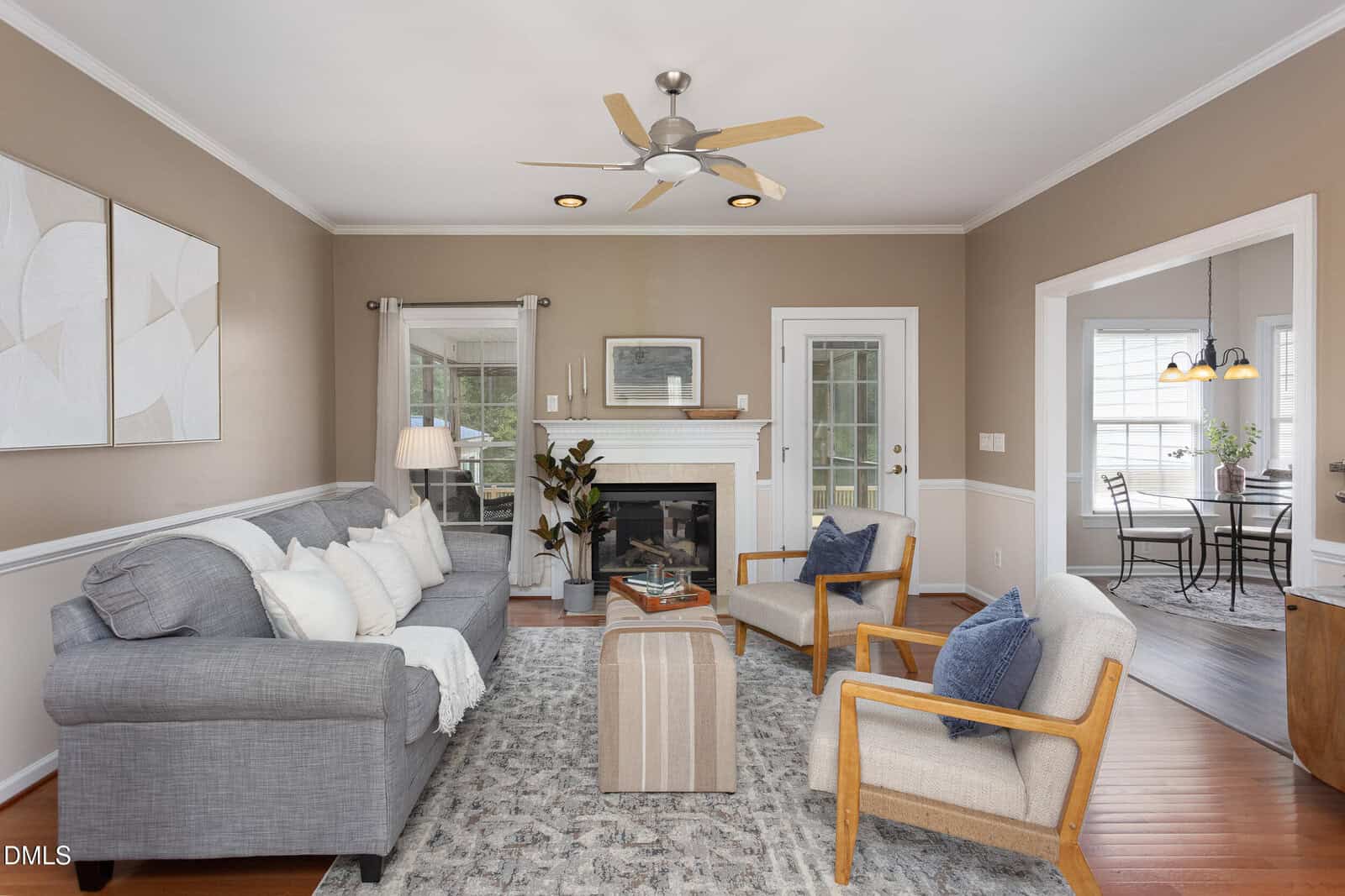 A living room with a gray sofa, two beige armchairs, a patterned rug, a fireplace, and a ceiling fan. Neutral walls and modern decor. A dining area is visible through an open doorway on the right.