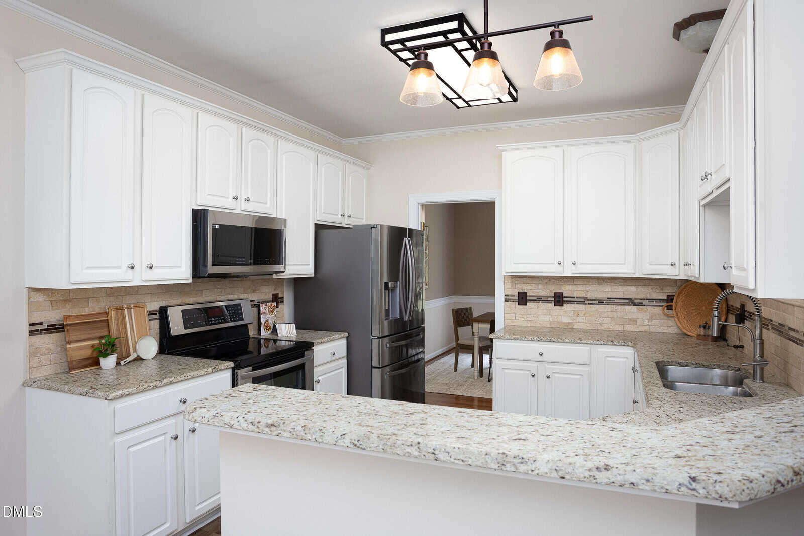 Modern kitchen with white cabinets, stainless steel appliances, beige granite countertops, tiled backsplash, and a three-light fixture over the bar. A small plant and cutting boards sit on the counter.