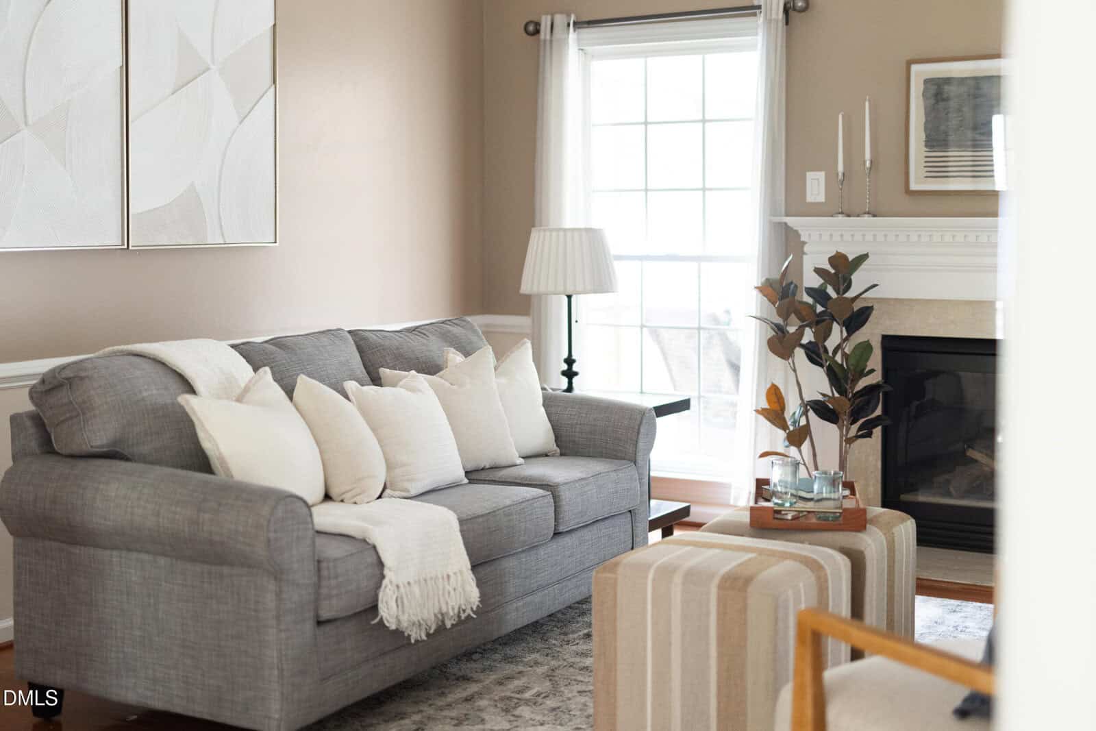 A living room with a gray sofa, white and beige pillows, a throw blanket, a striped ottoman, a small plant, a lamp, and a fireplace with candles and decorative objects on the mantel. Light streams in from a window.