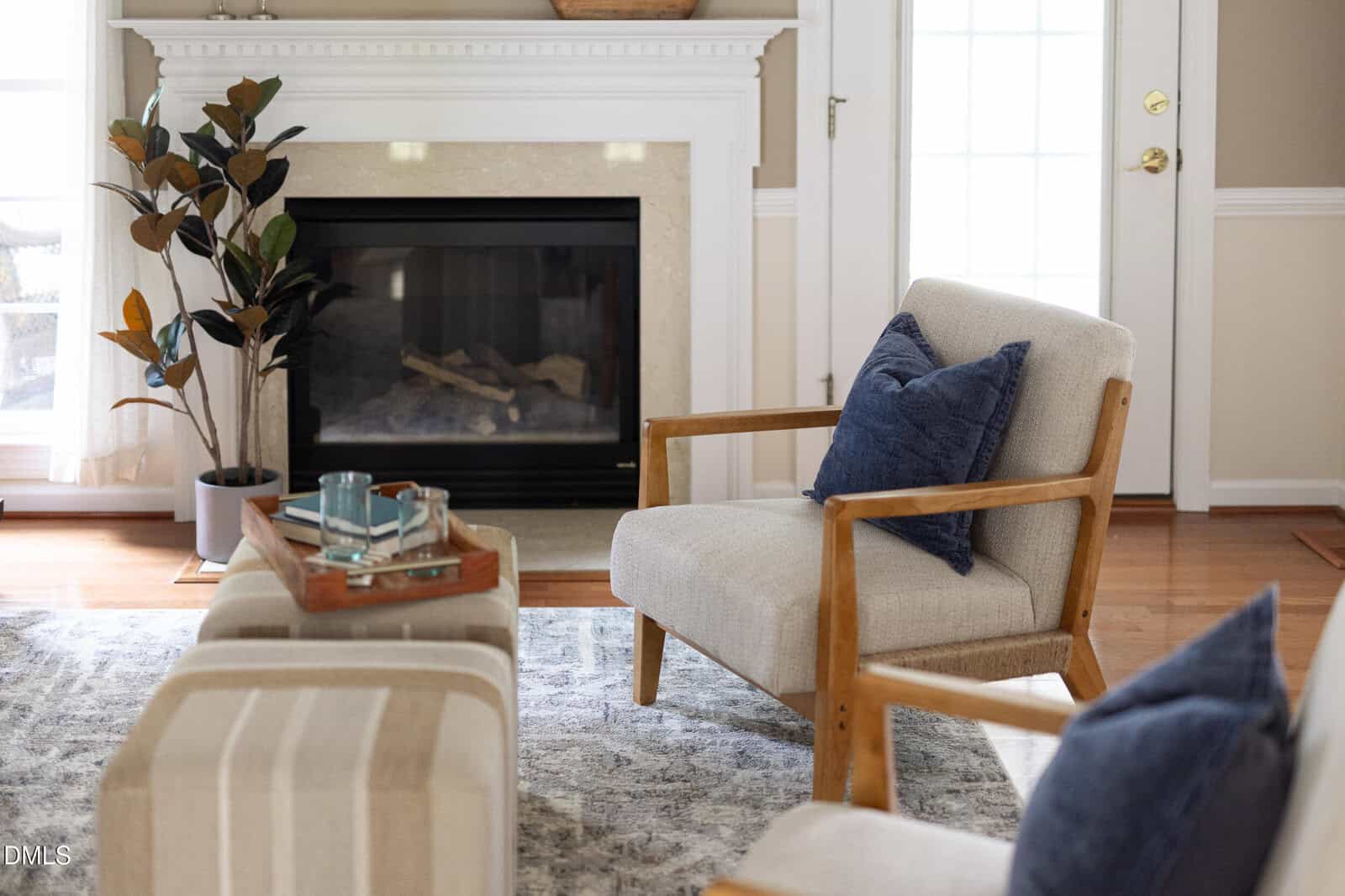 A cozy living room with a fireplace, armchair with blue pillow, striped ottoman, indoor plant, and a tray with cups on a soft rug. Sunlight filters through a window and glass door.
