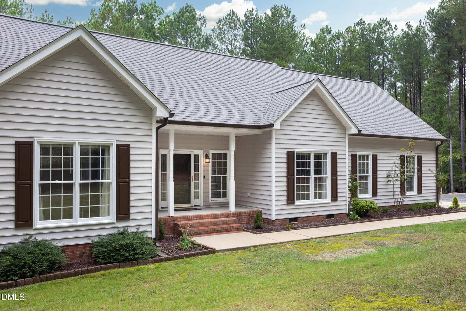 Single-story home with light gray siding, white trim, dark shutters, a covered front porch, brick steps, and multiple large windows. The house is surrounded by trees and has a sidewalk and landscaped front yard.