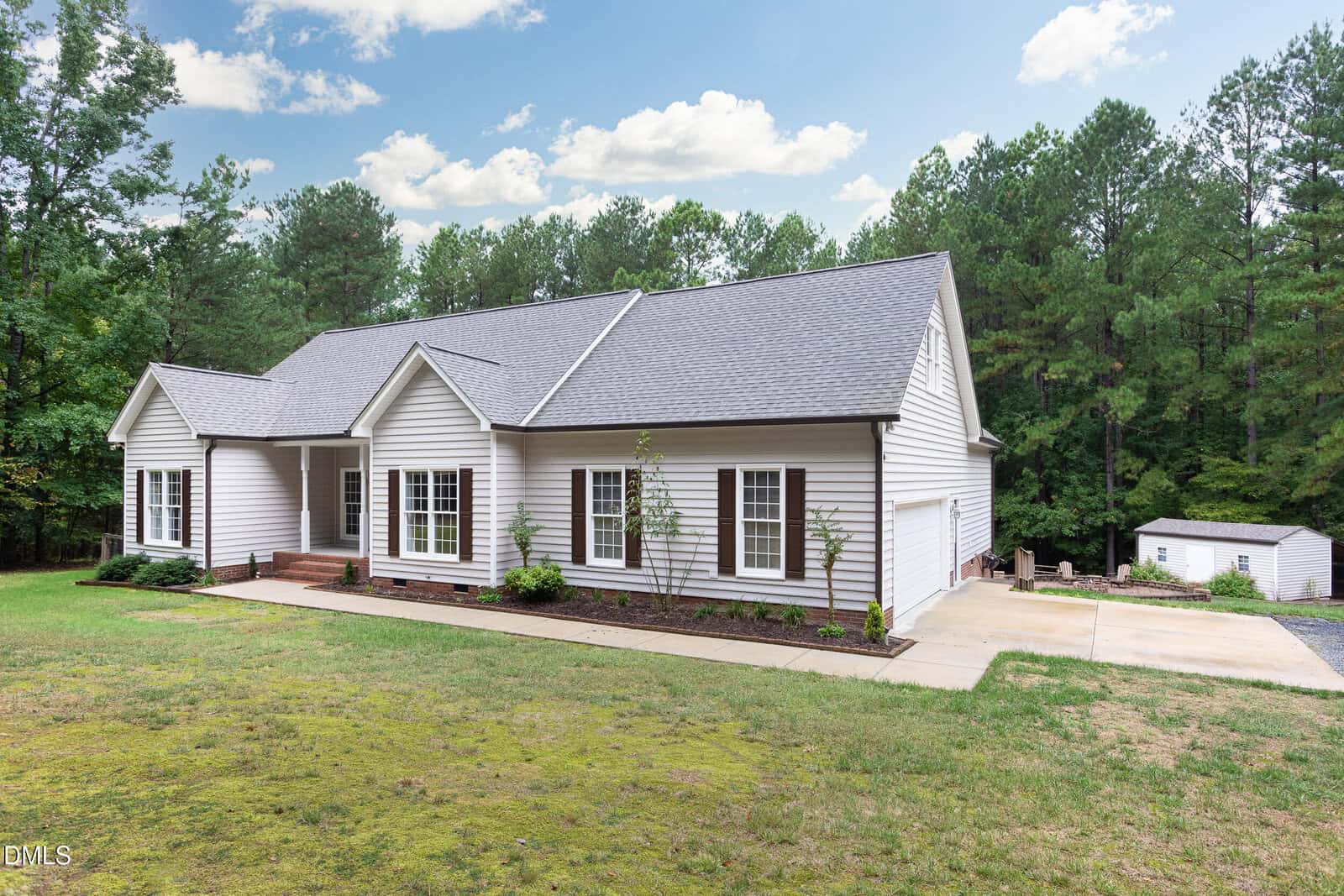 A single-story white house with a gray roof and attached porch, surrounded by trees. There is a concrete driveway leading to a detached garage in the background, with a grassy lawn in front.