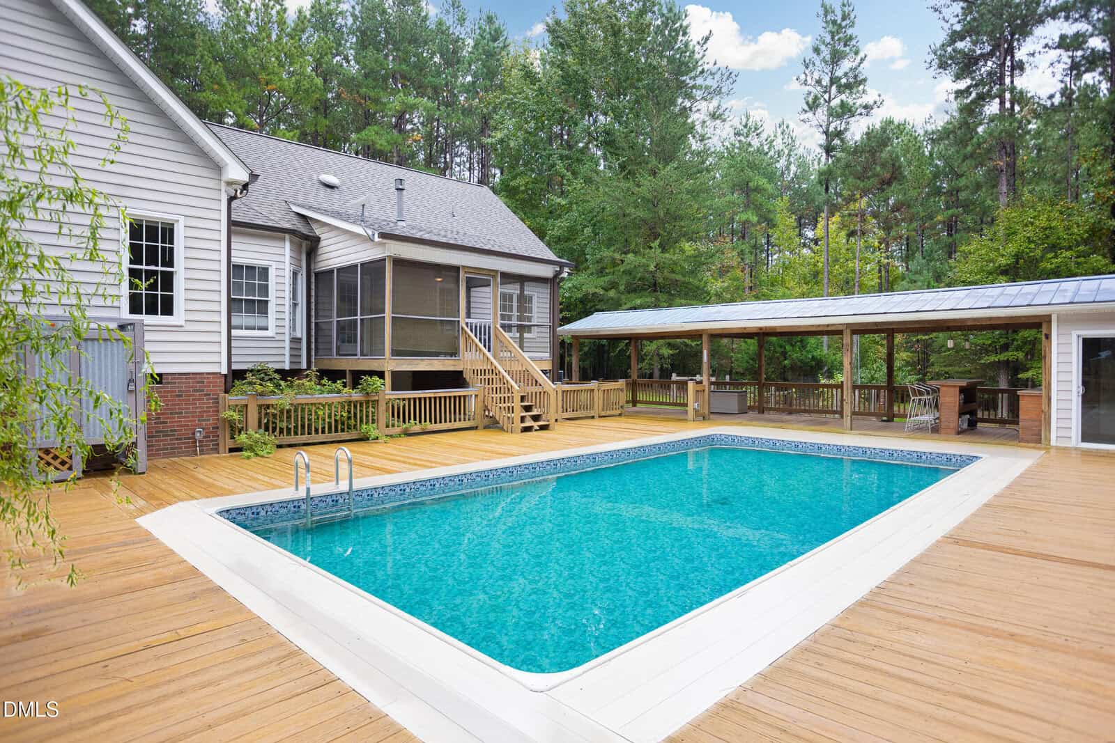 A rectangular in-ground swimming pool surrounded by a wooden deck is in the backyard of a house. The house has gray siding, a screened-in porch, and is bordered by trees and greenery.