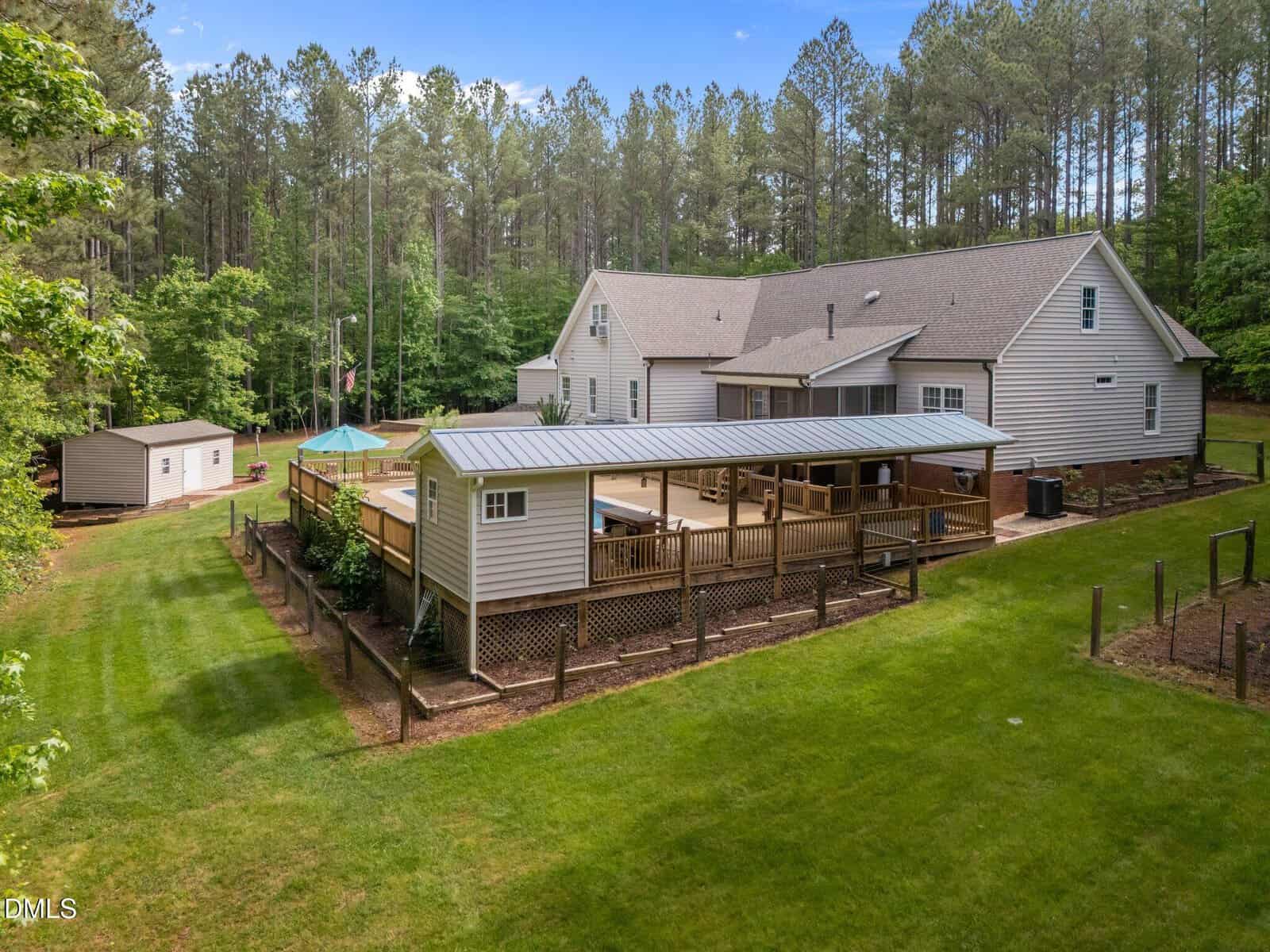 A large light-colored house with a sloped roof, a covered deck, and an above-ground pool in a spacious backyard surrounded by grass, trees, and a forested area. A shed is visible in the background.