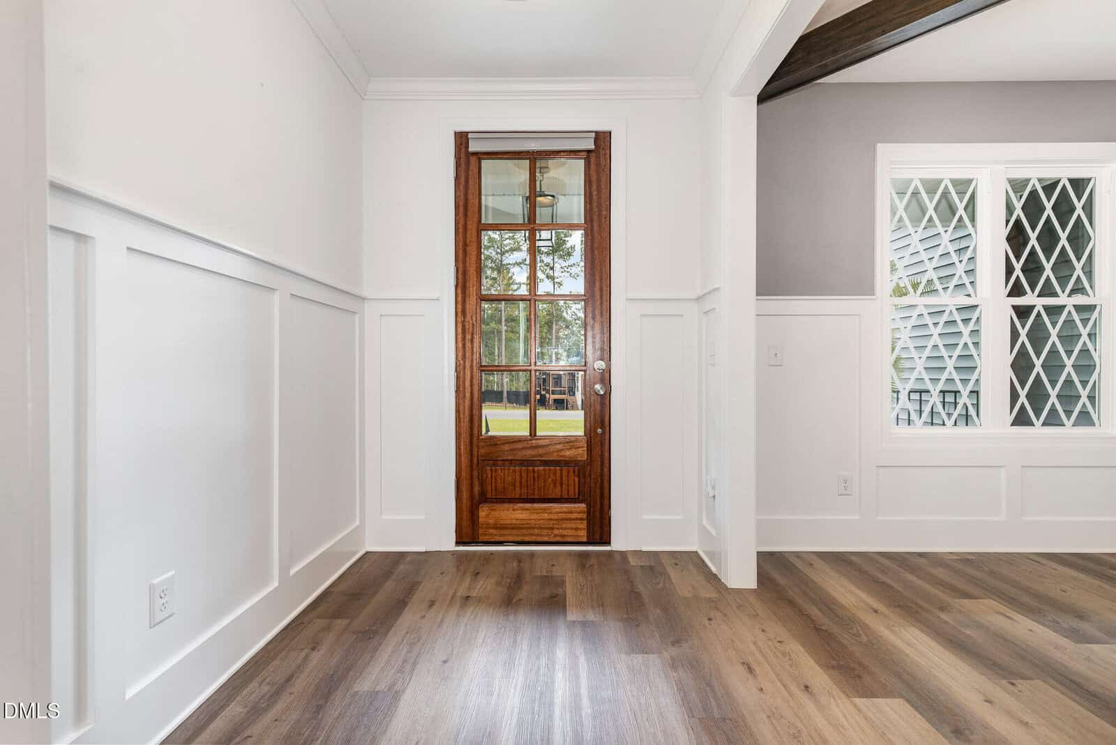 A room with white wainscoting, light wood floors, a wooden door with glass panes, and a window with a diamond-patterned grid. Outdoor trees are visible through the door.