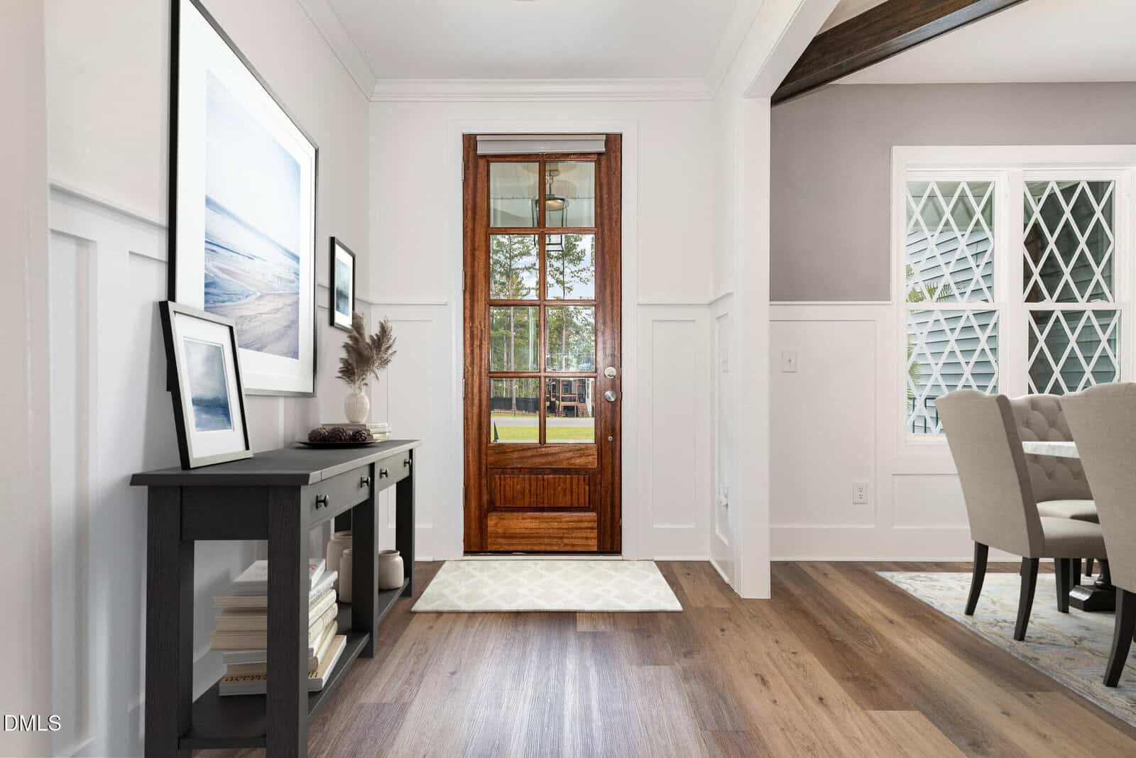 Entryway with a wooden door featuring glass panels, console table with decor and books, framed art, light wood flooring, view into dining area with upholstered chairs, and natural light from windows.