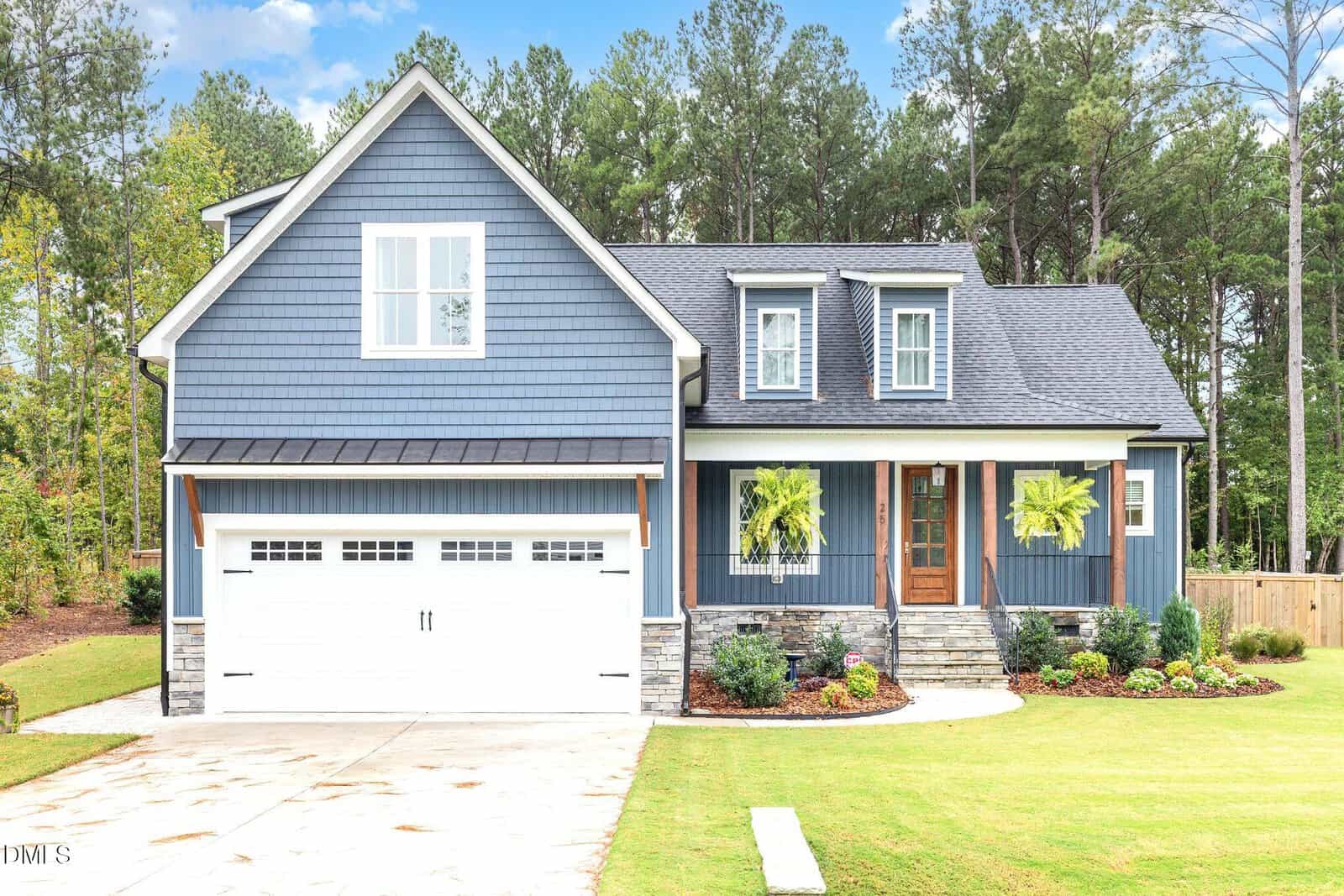 A blue two-story suburban house with white trim, a double garage, front porch with columns, two hanging ferns, stone steps, and a manicured lawn with shrubs sits gracefully at 25 Daisy Drive in Franklinton, surrounded by tall trees in the background.