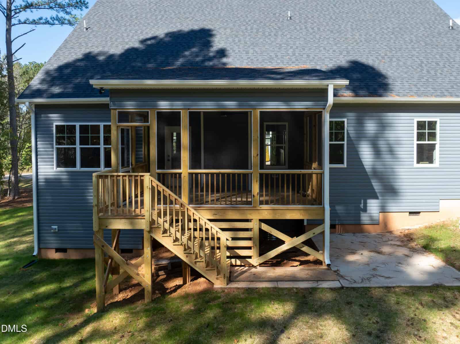 The rear view of a blue-sided house with a raised wooden screened-in porch. A staircase leads down from the porch to a concrete patio in the backyard. Shadows from nearby trees are cast across the roof and the siding of the house.