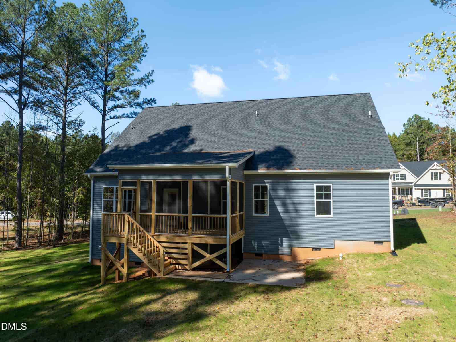 A blue house with a dark roof features a screened-in back porch with wooden stairs and railing, set on a grassy yard bordered by tall trees under a clear sky.